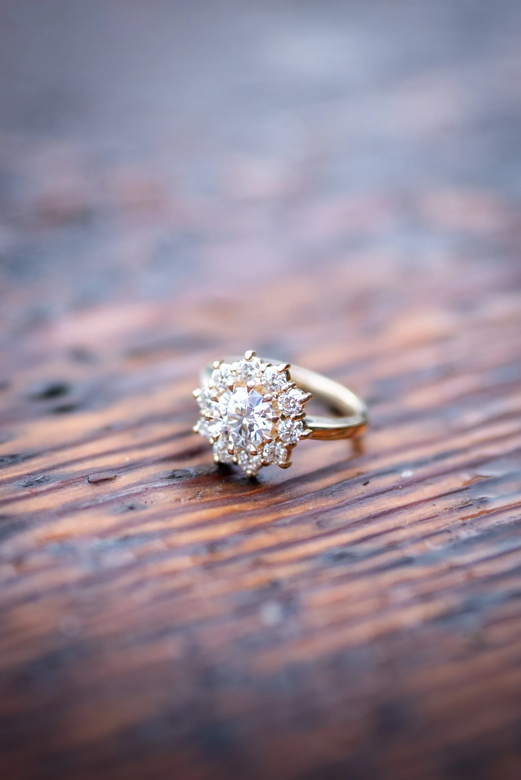 A diamond engagement ring with a large central stone surrounded by smaller stones, resting on a textured wooden surface with a blurred, colorful background.