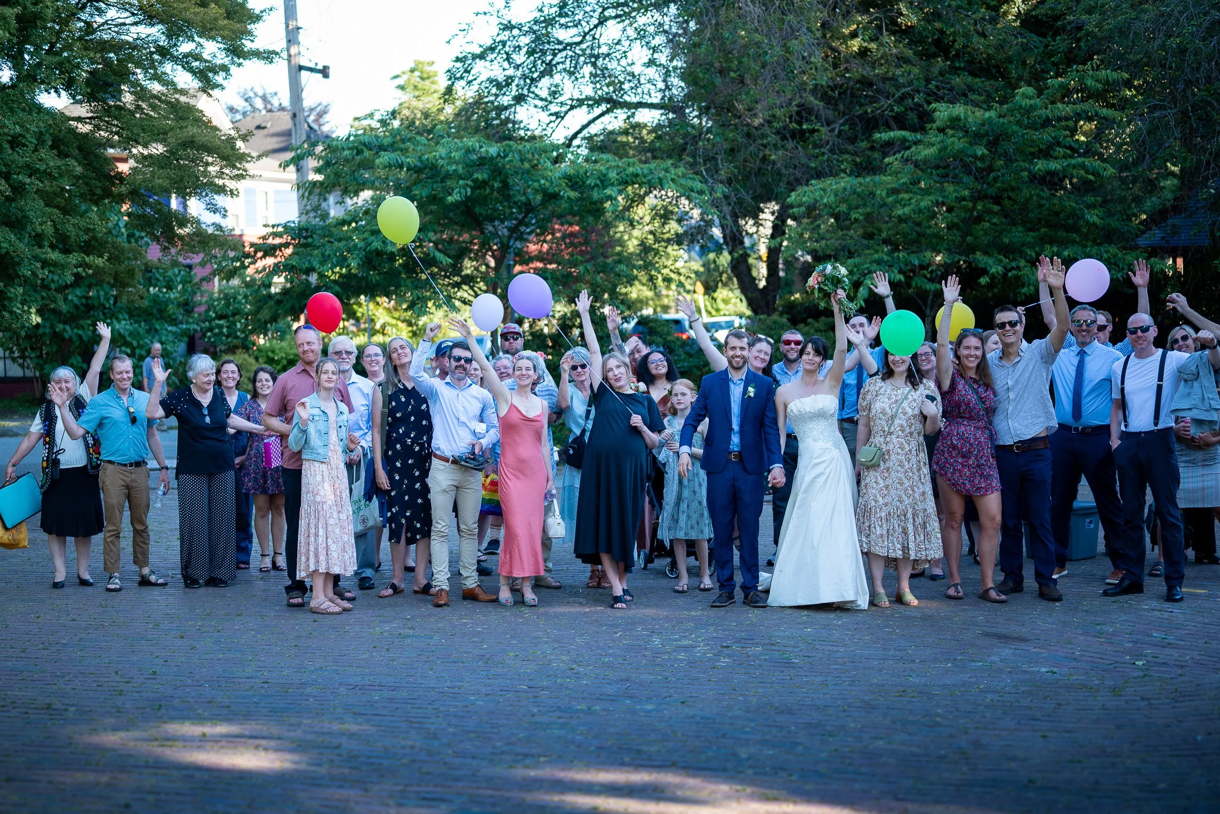 A diverse group of people, including a bride in a white wedding dress, are gathered outdoors on a sunny day, celebrating and smiling. Some are holding colorful balloons, and others are raising their hands. The setting is a park with green trees and b