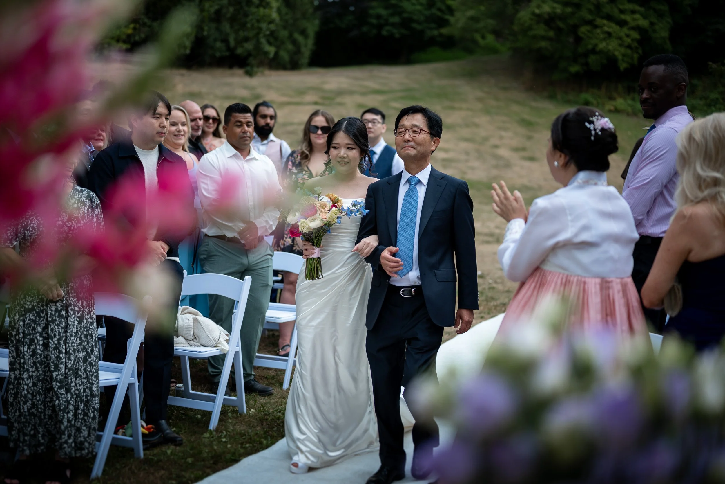 A bride in a wedding dress holding a bouquet walking down an outdoor aisle with her father, surrounded by wedding guests seated and standing on a grassy area, with trees in the background.