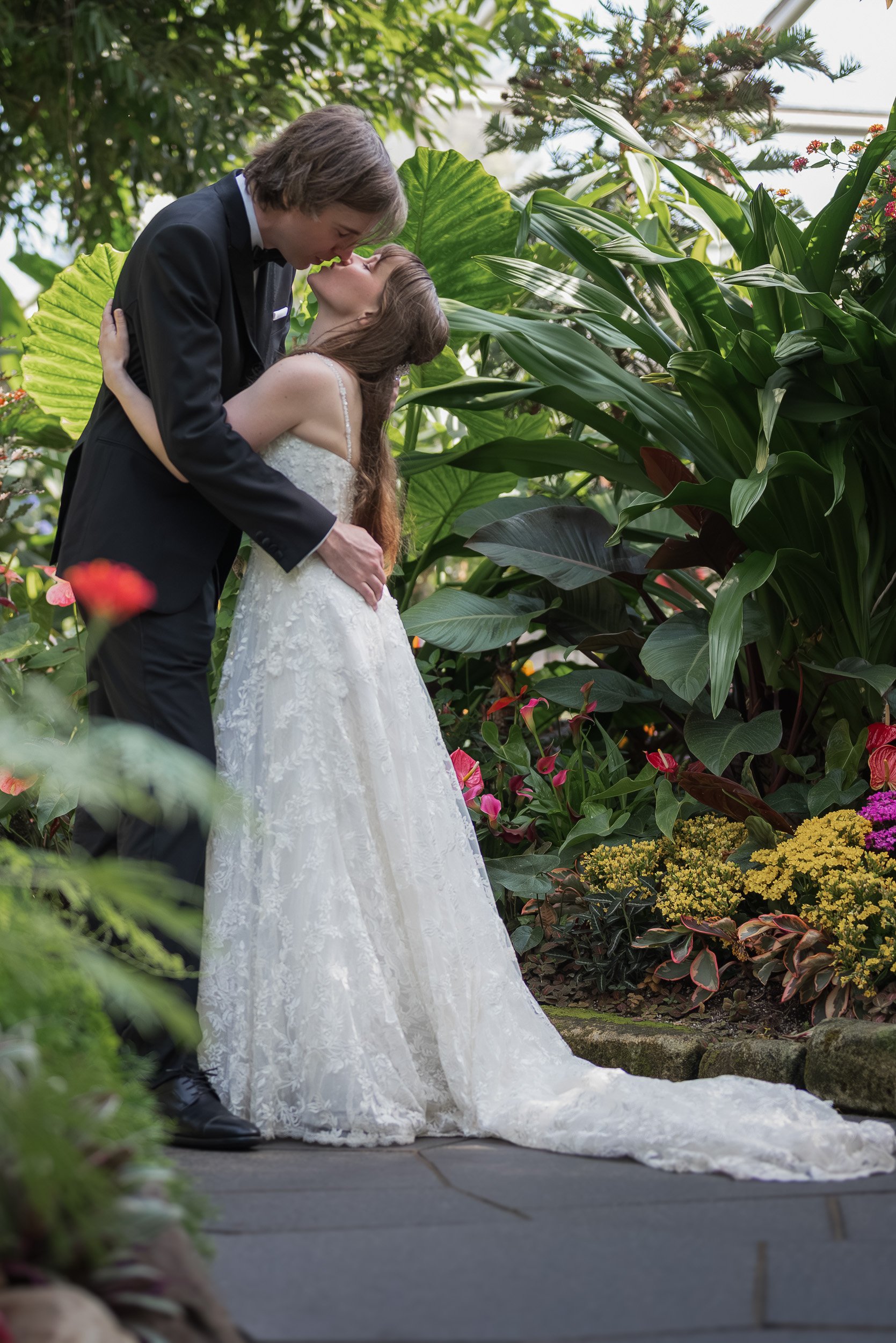 A bride and groom in wedding attire at Bloedel Conservatory sharing a kiss in a lush garden filled with large green leaves and colorful flowers.