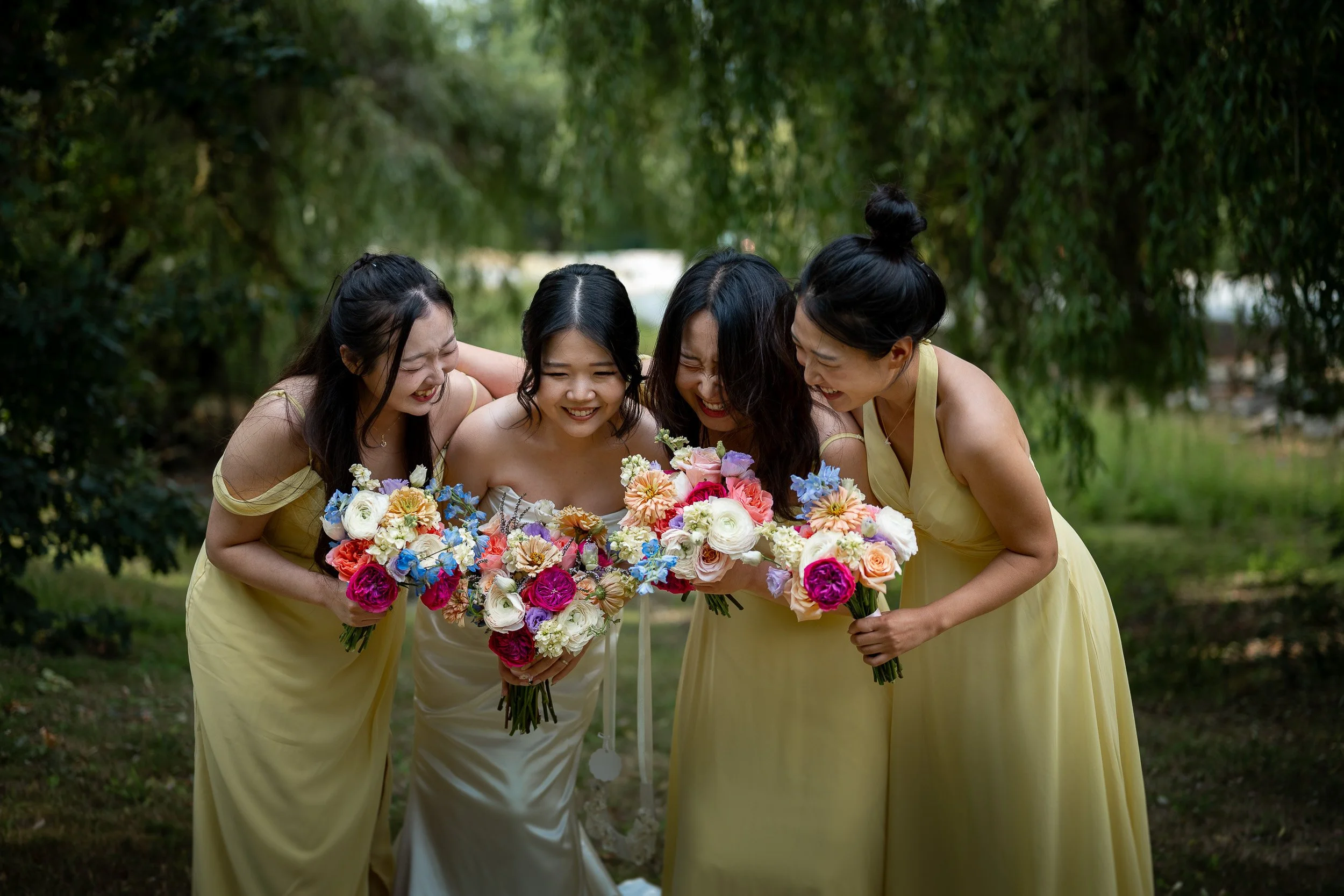 Four women in yellow dresses smiling and holding bouquets of colorful flowers outdoors, surrounded by green trees.
