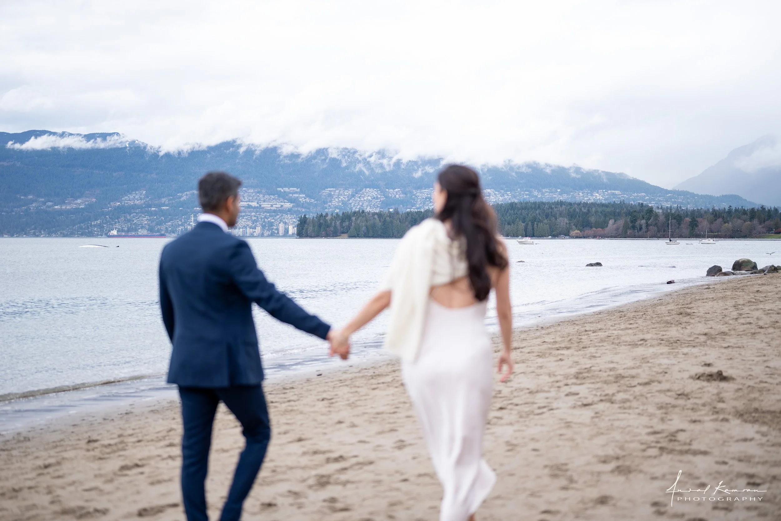 A couple in wedding attire holding hands and walking along a beach with mountains and cloudy sky in the background at their wedding at Kitsilano Beach