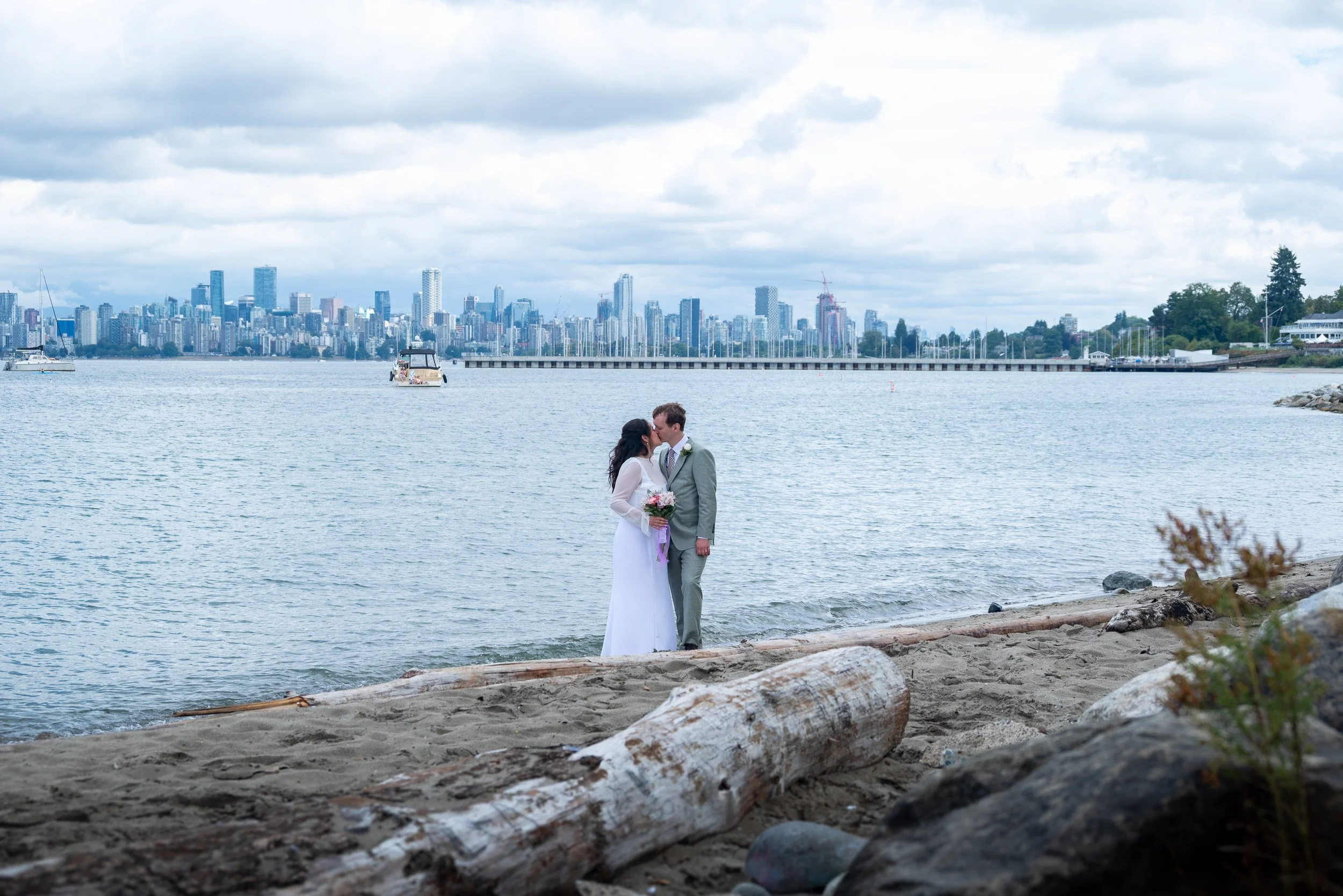 A bride and groom kissing by the ocean at Jericho Beach in Vancouver with a Vancouver city skyline in the background, boats on the water, and cloudy sky.