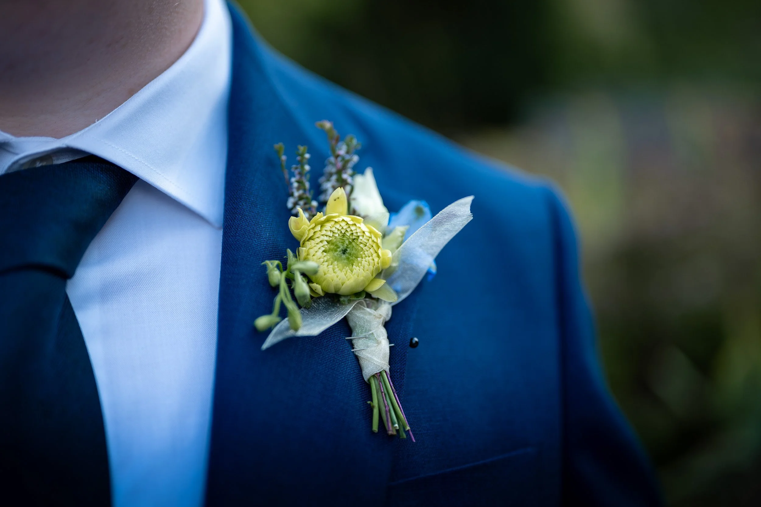 Close-up of a man in a blue suit and white shirt wearing a boutonniere with a yellow flower and greenery.