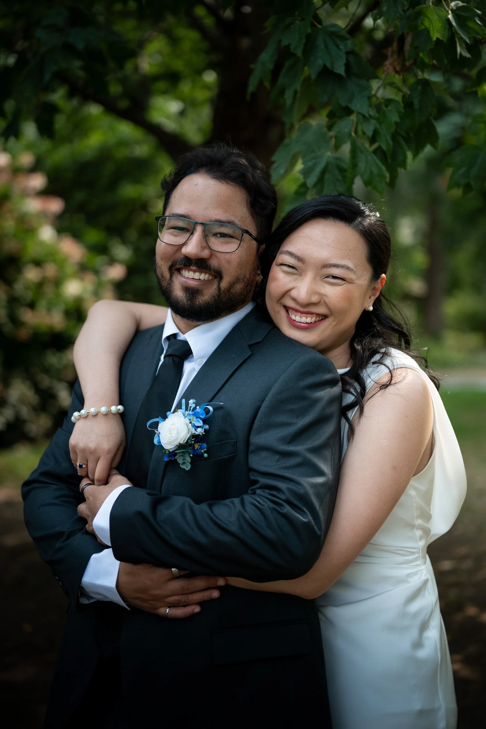 A happy couple dressed in formal attire, smiling and hugging outdoors in a green park with trees in the background.
