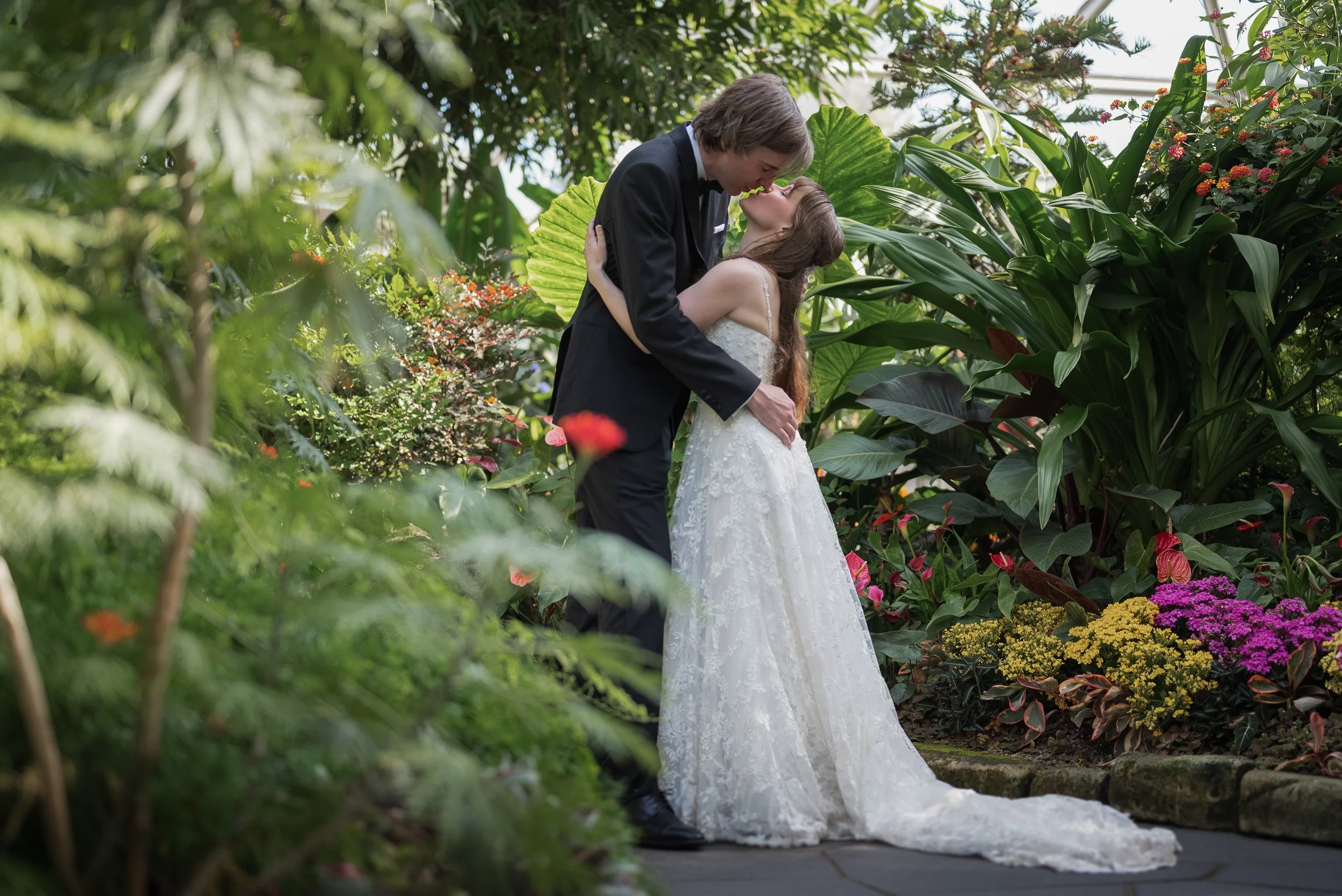 A couple in wedding attire standing close together at Bloedel Conservatory, sharing a romantic moment surrounded by vibrant green plants and colorful flowers.