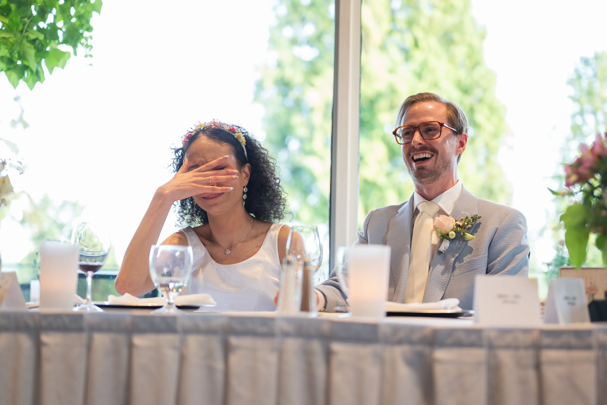 A bride and groom sitting at a wedding reception table, with the bride covering her face with her hand and the groom smiling, in a room with large windows and green trees outside.