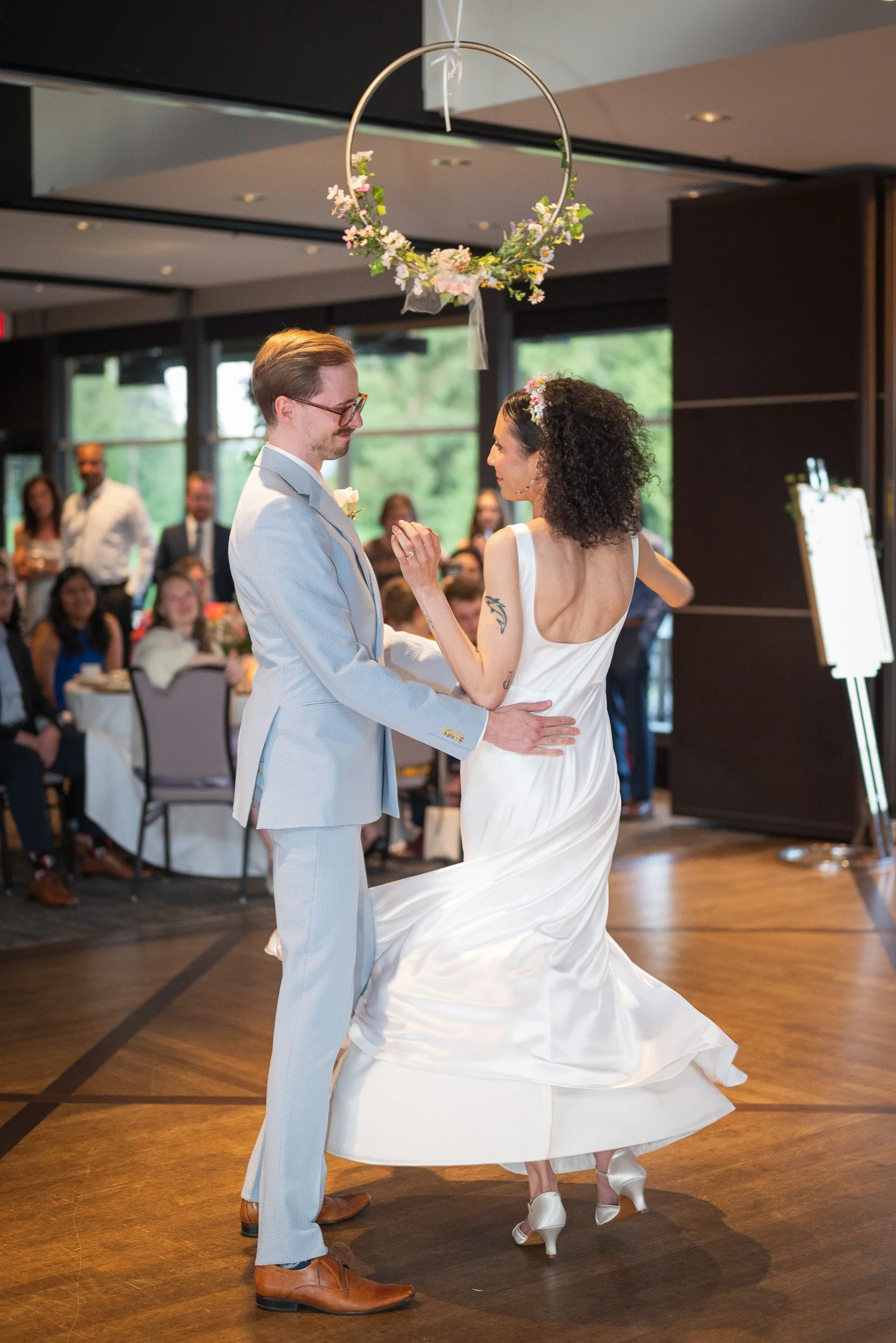A couple is dancing during their wedding reception, with the bride in a white dress and the groom in a light gray suit, in a decorated indoor venue with guests watching in the background.
