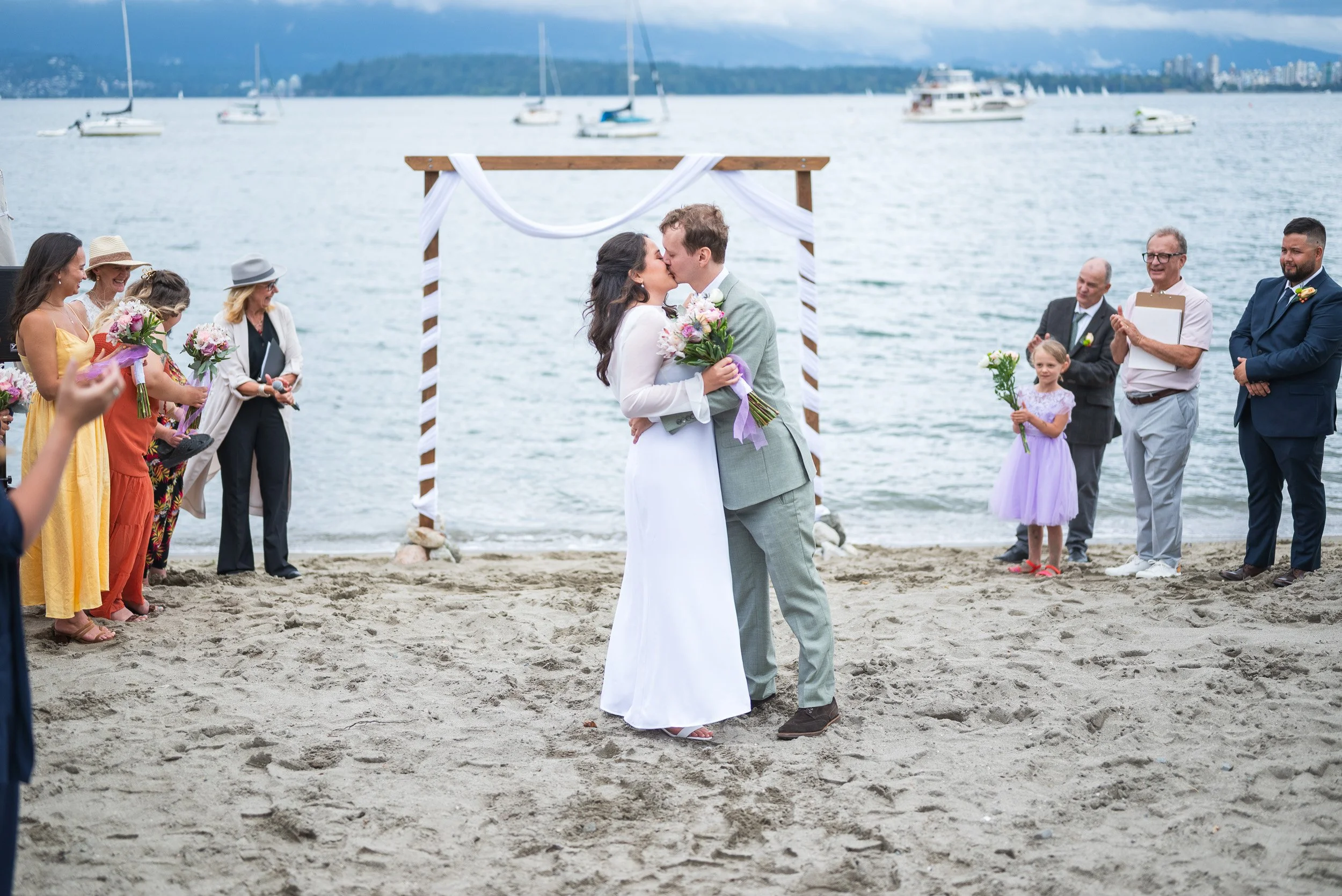A wedding ceremony taking place on Jericho Beach with the ocean and sailboats in the background. The bride and groom are kissing under a wooden arch decorated with white fabric. Guests are standing on the sandy shore, holding flowers and smiling.
