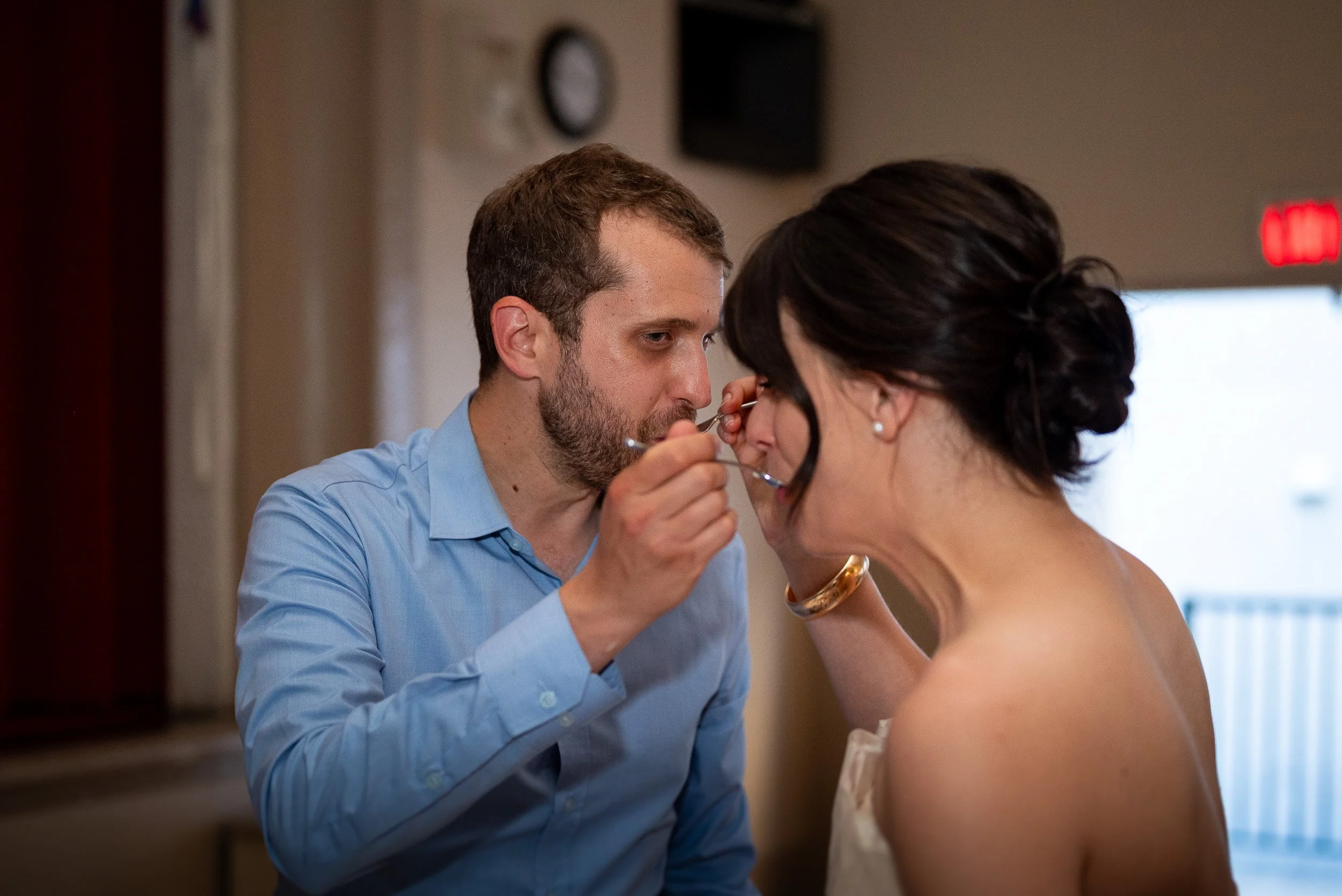 A man in a blue shirt is applying makeup or grooming a woman's face, who is wearing glasses and a white dress, inside a room with beige walls and a clock on the wall.