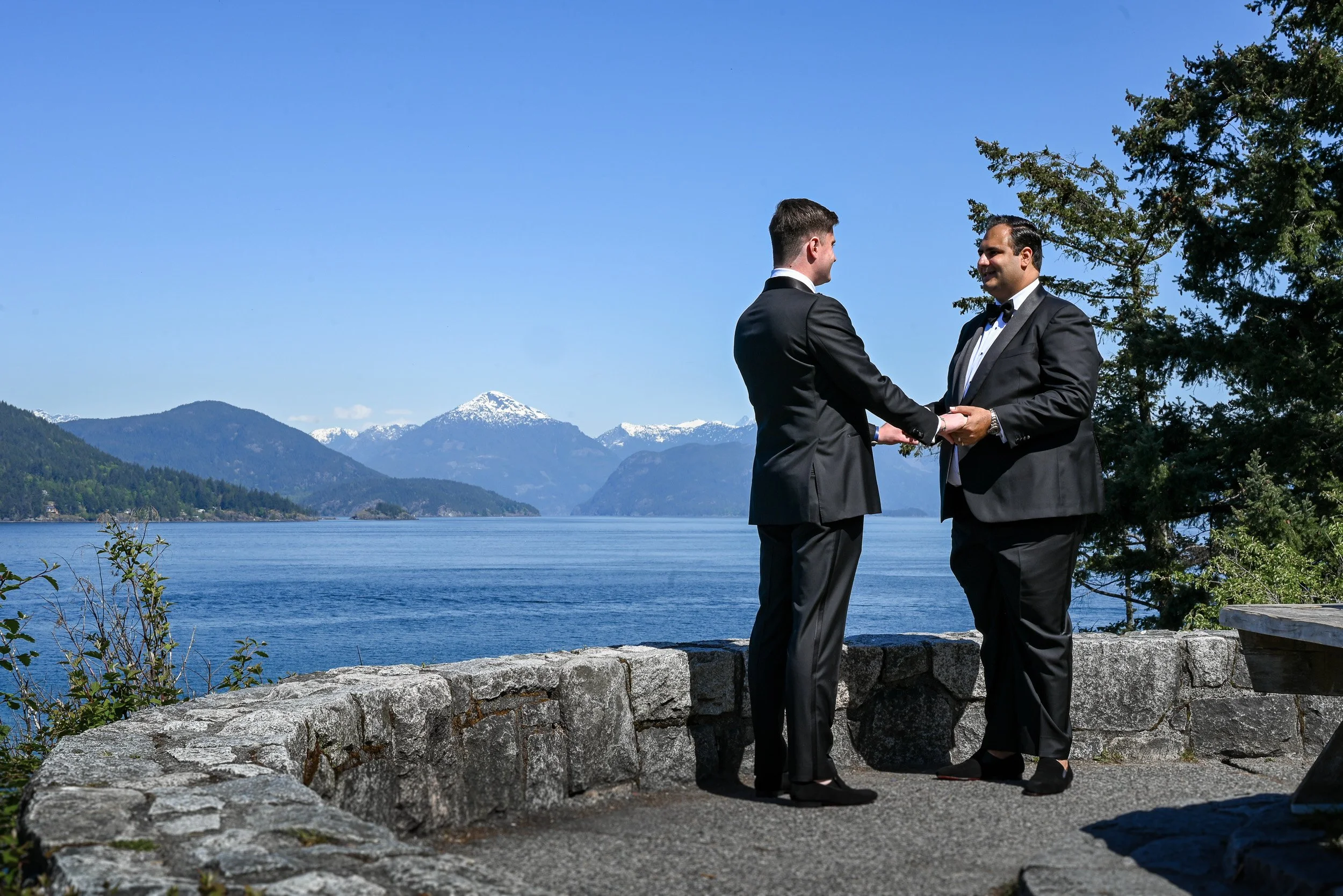 Two men in tuxedos exchanging wedding vows by a lake with mountain scenery in the background.