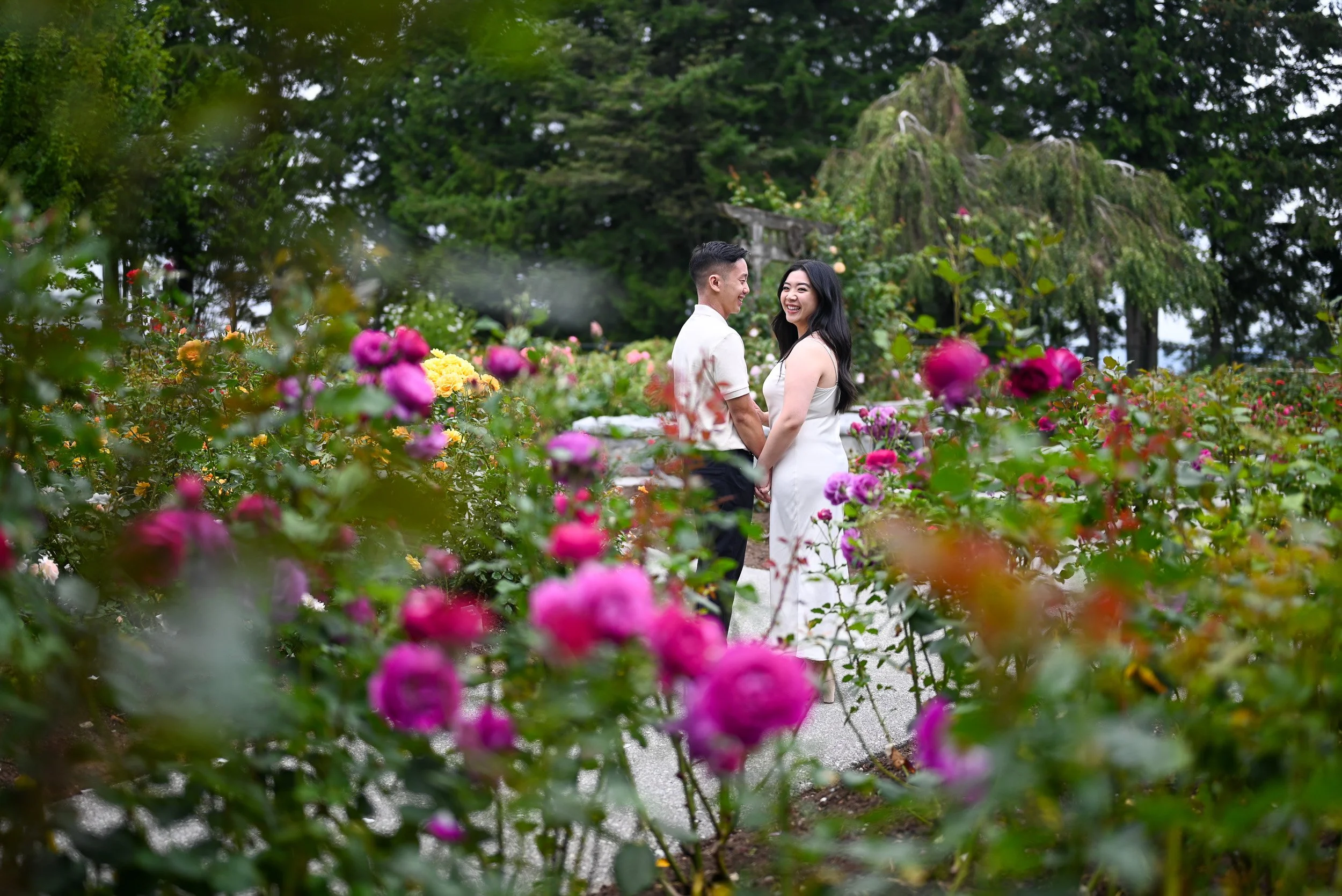 A couple holding hands and smiling at each other in a garden full of colorful pink and purple flowers at a Burnaby Park Elopement.