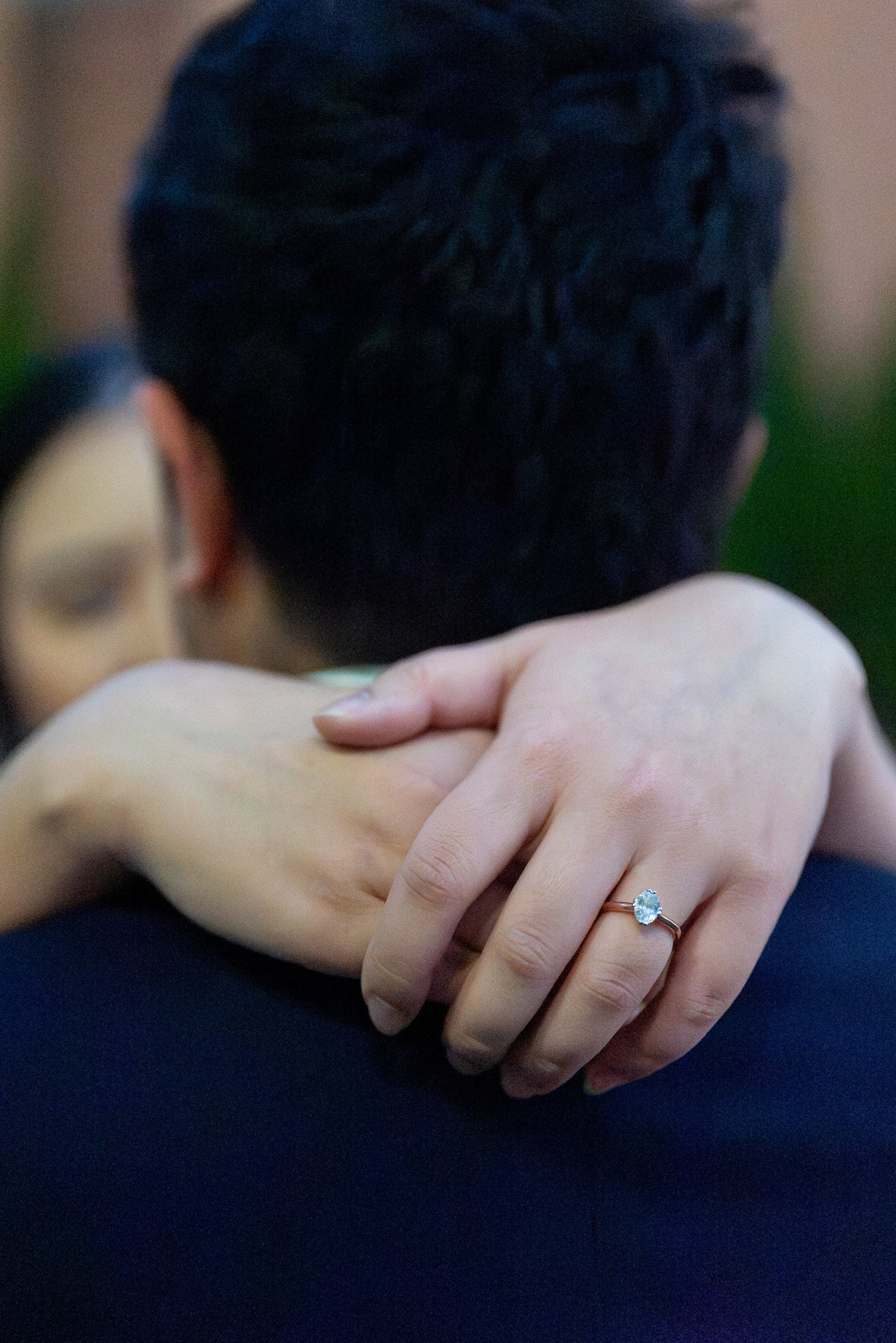 Close-up of a couple embracing, focusing on their hands. The woman's hand shows a ring with a large gemstone. The background is blurred, highlighting their intimate moment.