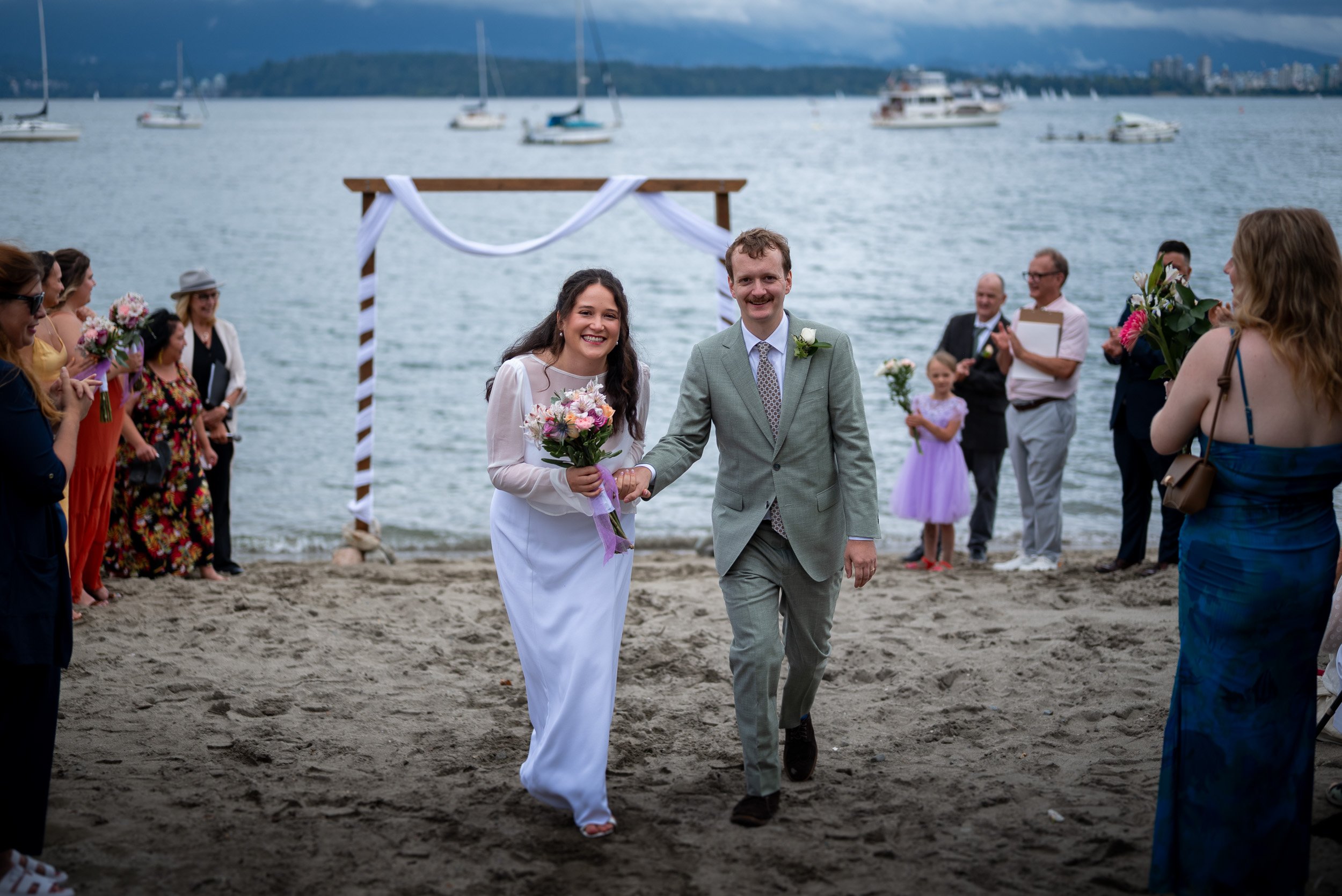 A couple, dressed in wedding attire, walking hand-in-hand on a beach during their wedding ceremony, with guests in the background and boats on the water.