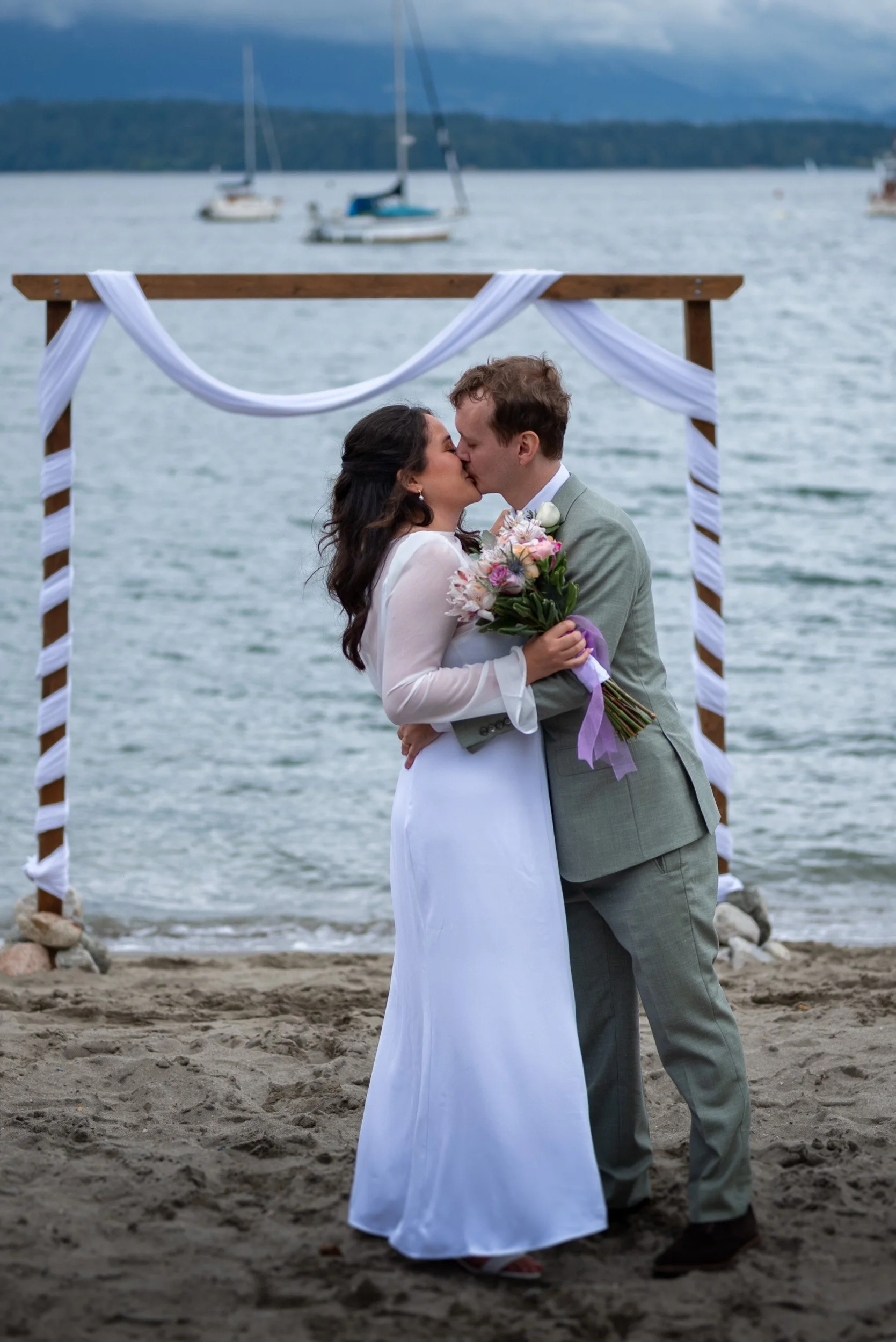 A bride and groom kissing during their wedding ceremony at Jericho Beach, with a wooden arch decorated with white fabric and boats in the water in the background.