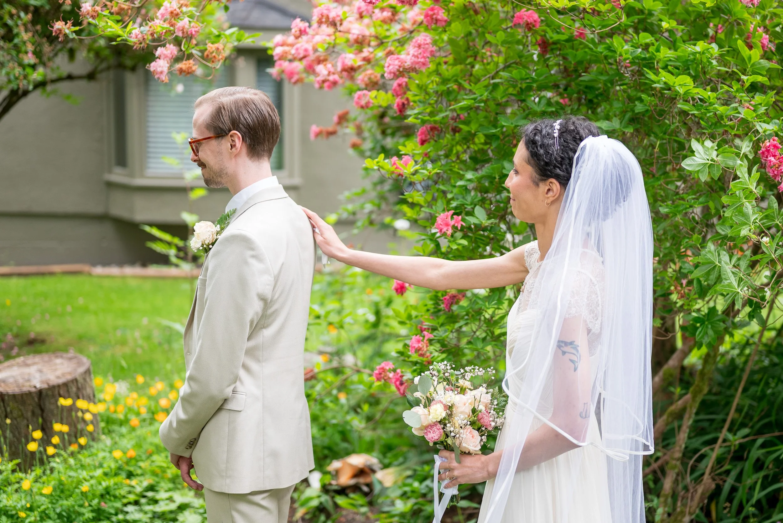 A newlywed couple stands in a garden surrounded by pink and green flowering bushes. The groom, dressed in a cream suit and glasses, faces away with his hands clasped. The bride, wearing a white lace wedding gown with a veil, gently touches the groom'