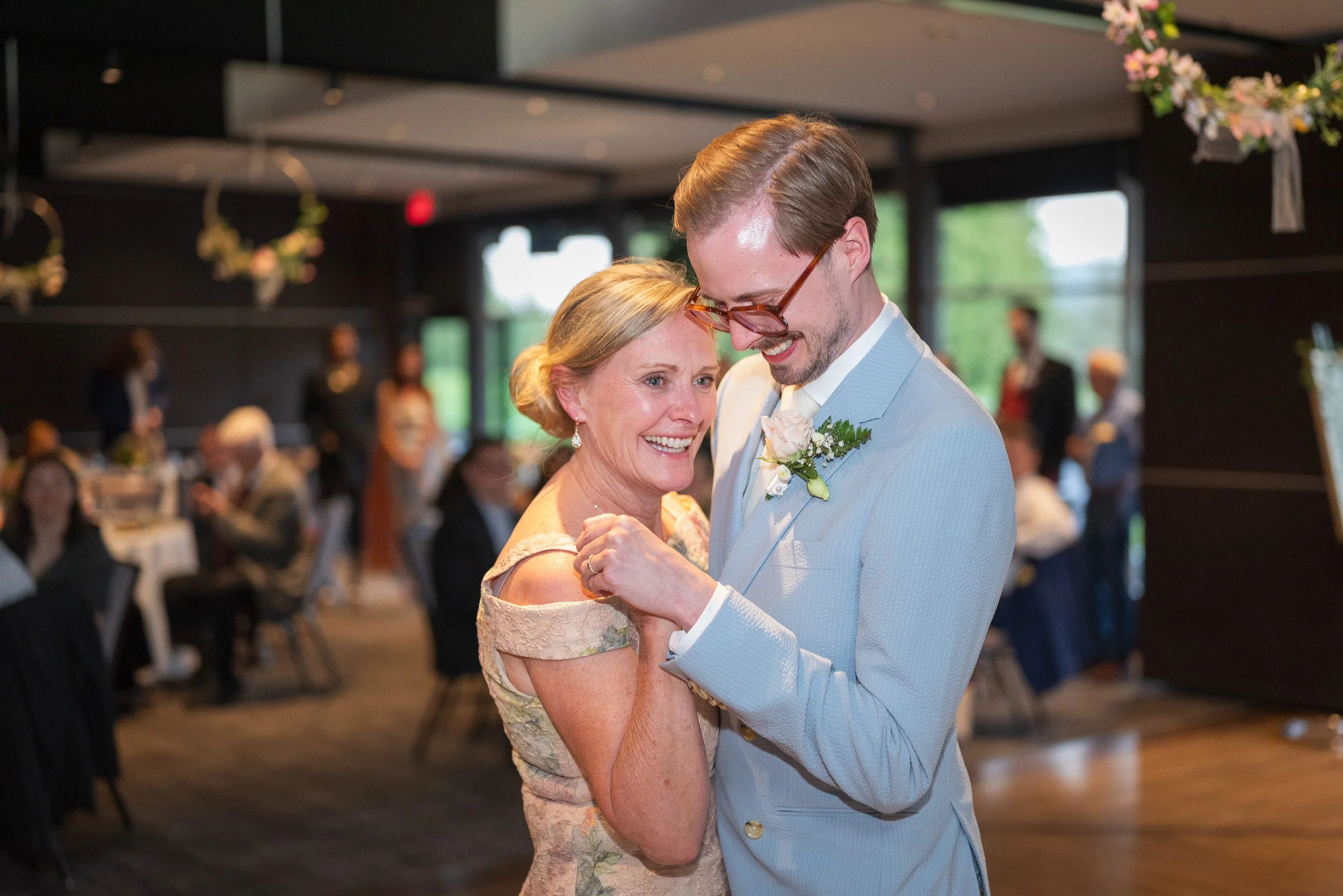 A young man and an older woman share a dance at a wedding reception. The young man is wearing a light blue suit and glasses, and the woman is in a floral dress. They are smiling and holding hands, with several guests seated at tables in the backgroun