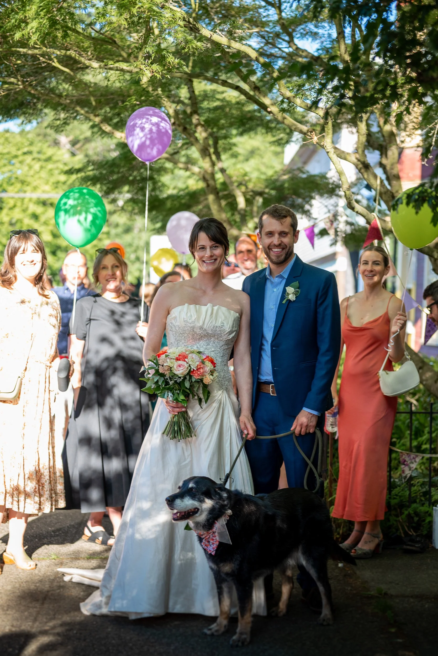 A bride and groom pose for a photo during their outdoor wedding celebration. The bride wears a strapless white gown and holds a bouquet of flowers, while the groom wears a blue suit with a boutonniere. They are surrounded by guests holding colorful b