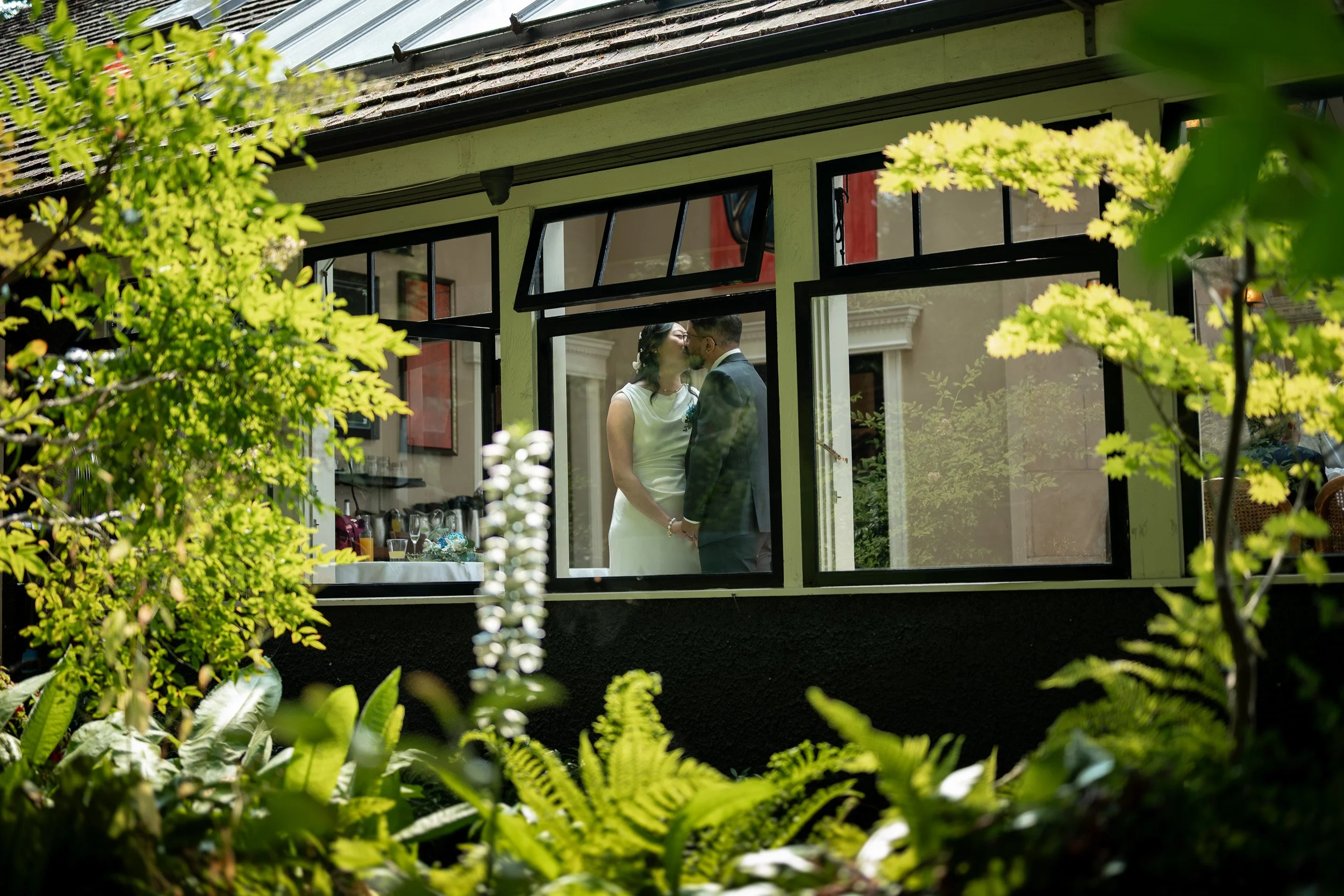 A couple kissing inside a house, viewed through open window from garden with green foliage.