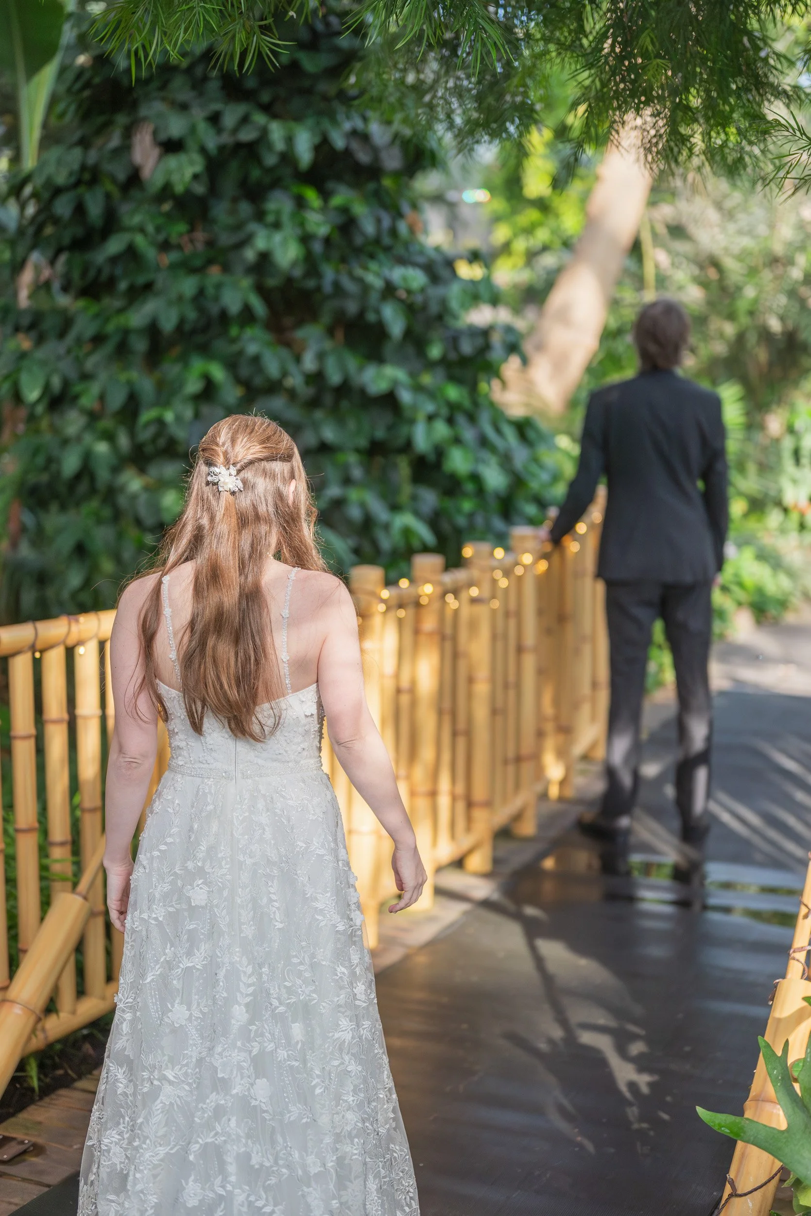 A woman in a white wedding dress walking on a wooden pathway surrounded by green foliage, with a man in a black suit ahead of her, walking away.