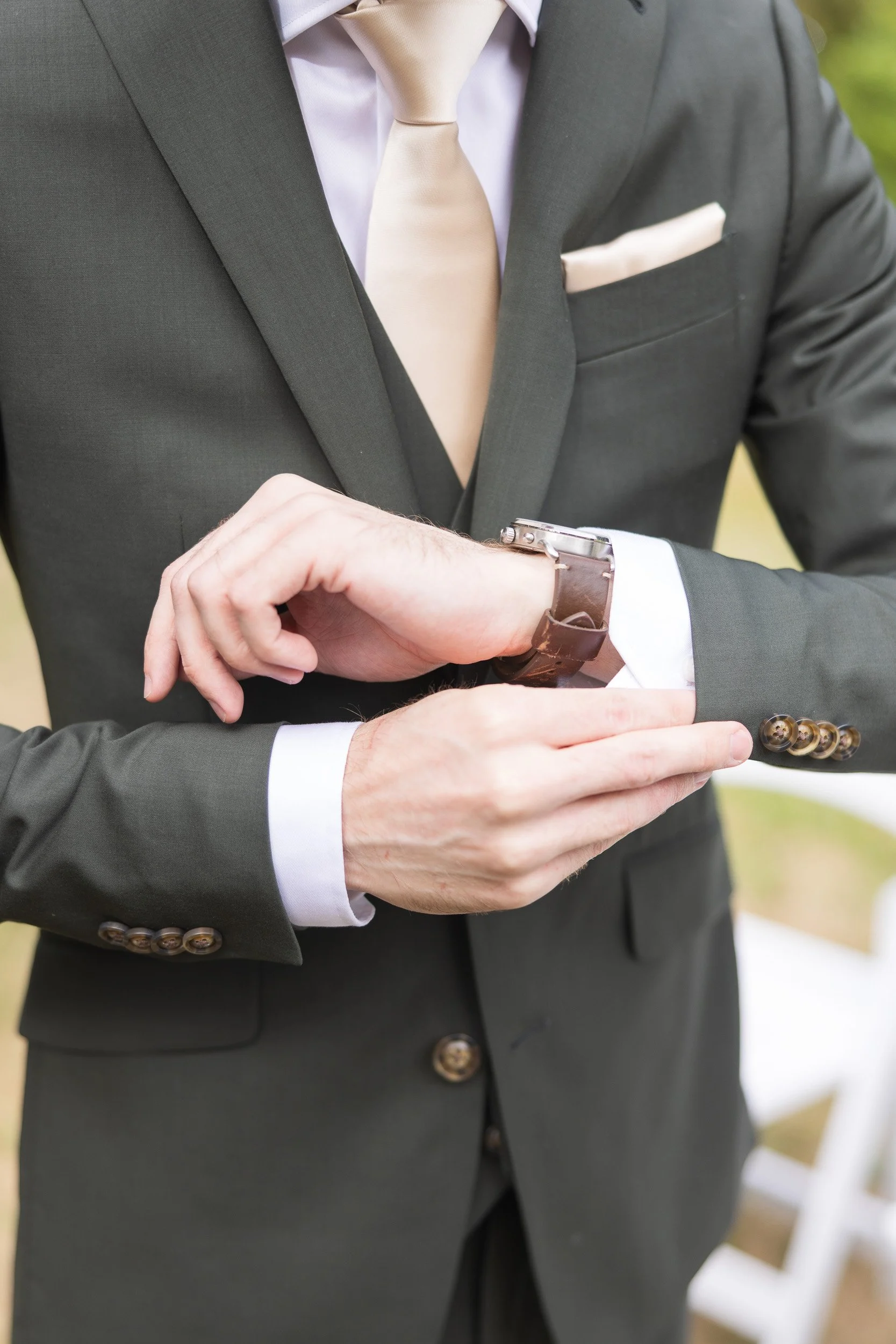 Close-up of a man dressed in a dark suit, white shirt, and beige tie, adjusting a wristwatch with a leather strap. He has a pocket square in his suit pocket and is outdoors with blurred greenery in the background.