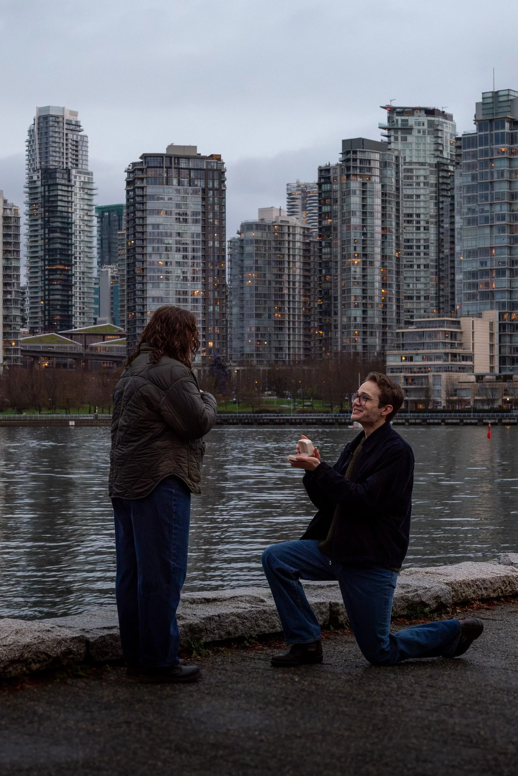 A man is kneeling on one knee while proposing to a woman in Vancouver with tall modern buildings in the background. The woman is standing with her hands crossed and the man is holding a small box, likely with an engagement ring inside.