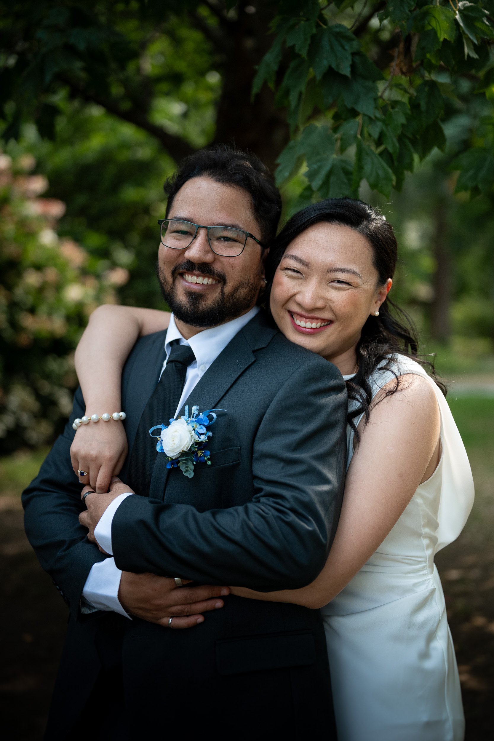 A smiling man in a tuxedo with glasses and a woman in a white dress with pearl earrings, embracing outdoors with green foliage in the background.