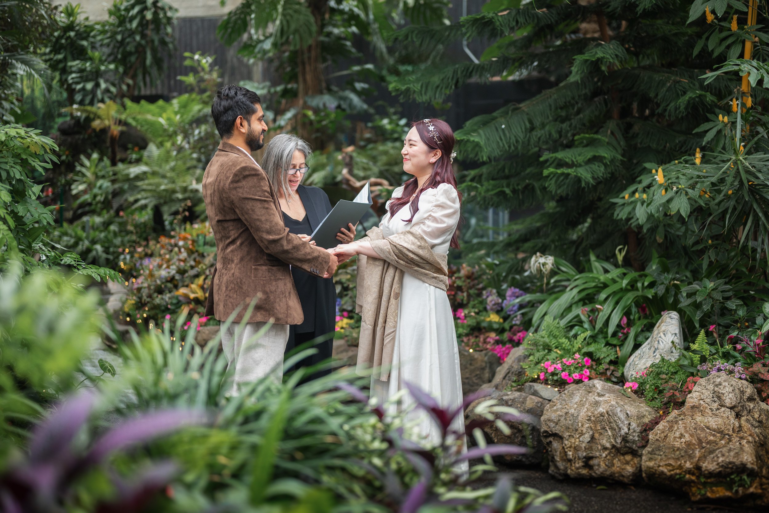 A couple and officiant during a wedding ceremony in Bloedel Conservatory, with the couple holding hands and smiling at each other.