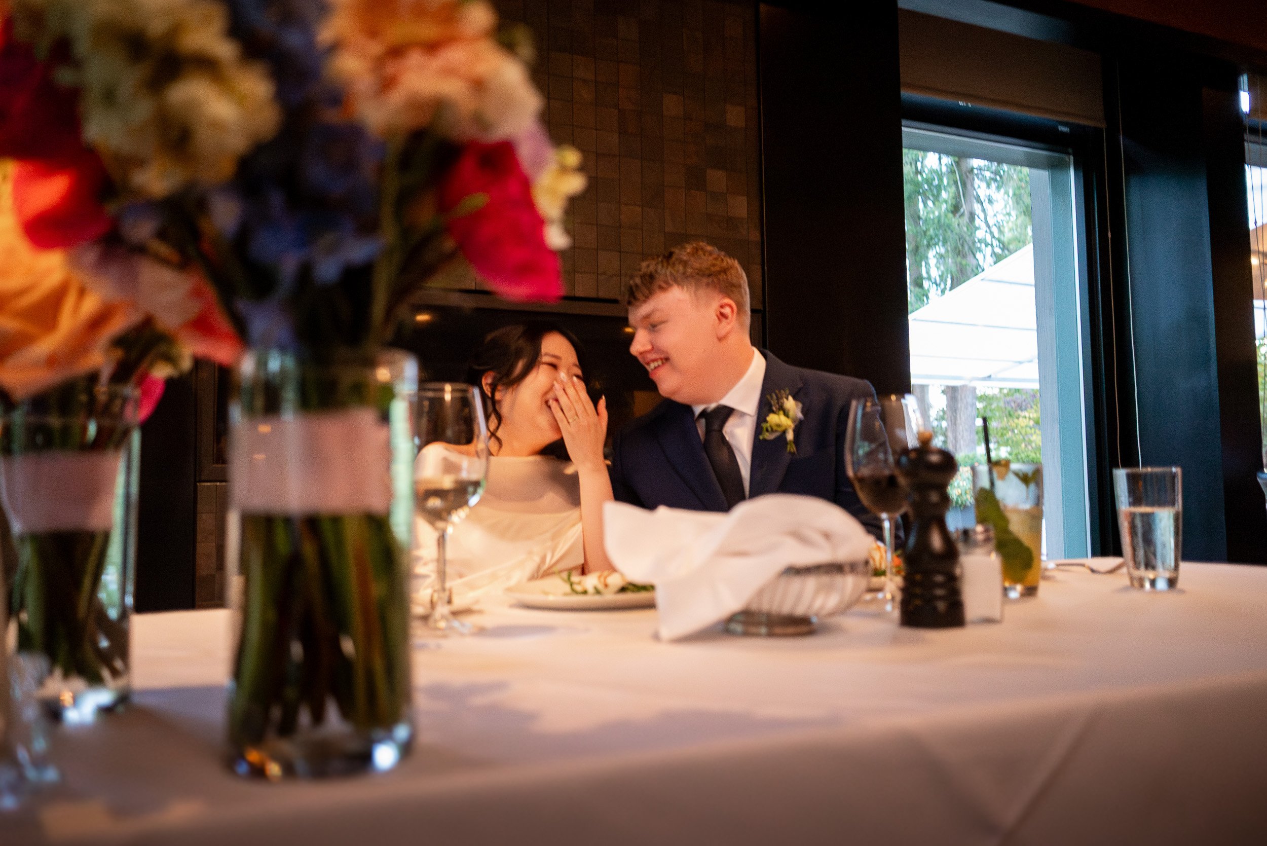 A happy bride and groom at a wedding reception at Seasons in the Park, sharing a joyful moment at the table. The focus is on the couple, with floral centerpieces in the foreground and a window behind them showing an outdoor scene.