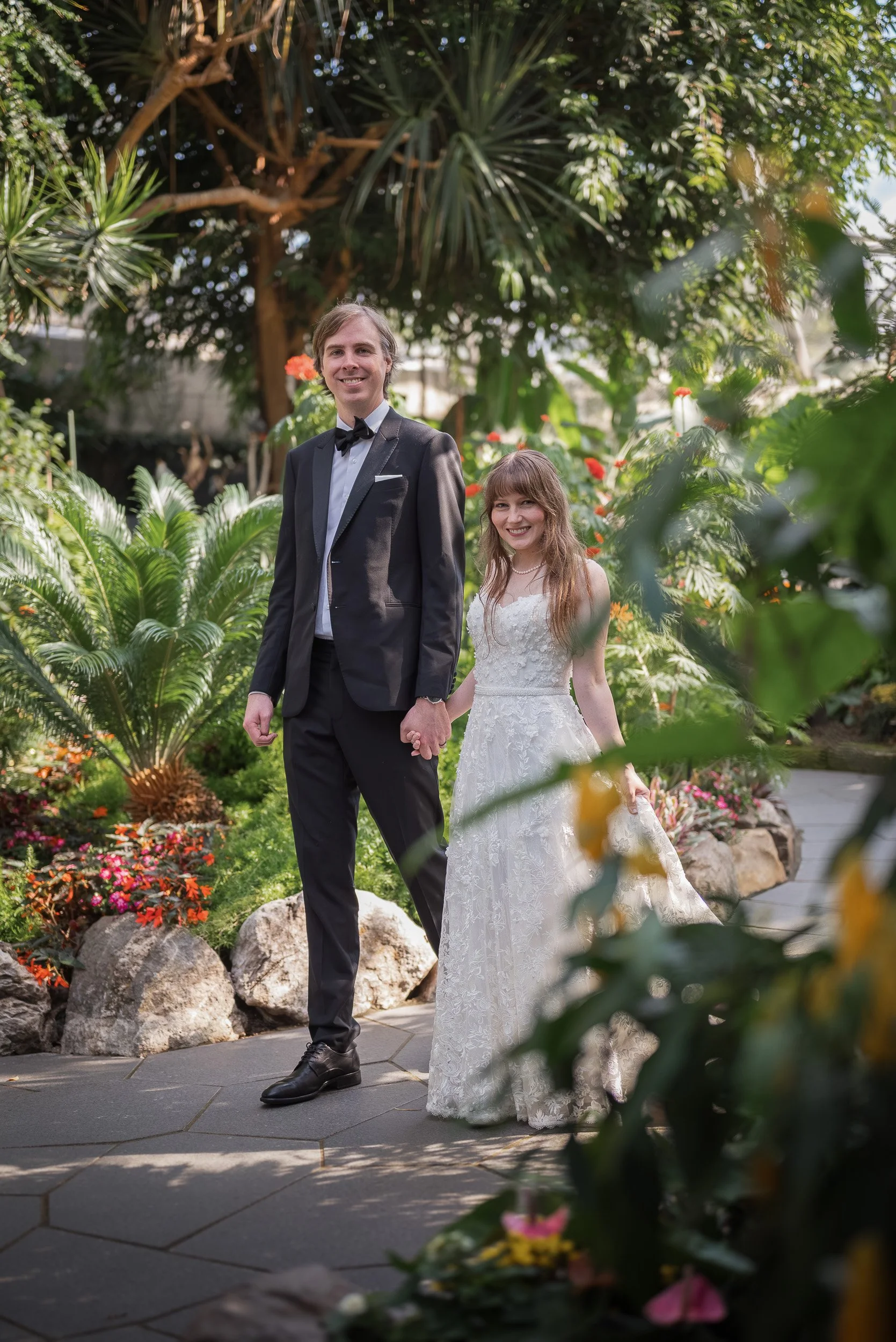 A young couple in wedding attire holding hands in a lush, green garden with various plants and flowers at Bloedel Conservatory.