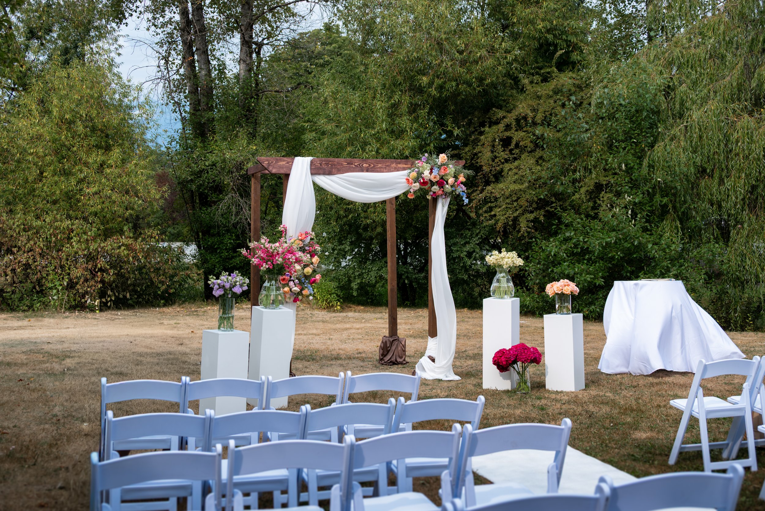 Outdoor wedding ceremony at Queen Elizabeth Park Duck Pond, setup with white chairs, a wooden arch decorated with white drapery and colorful flowers, surrounded by green trees.