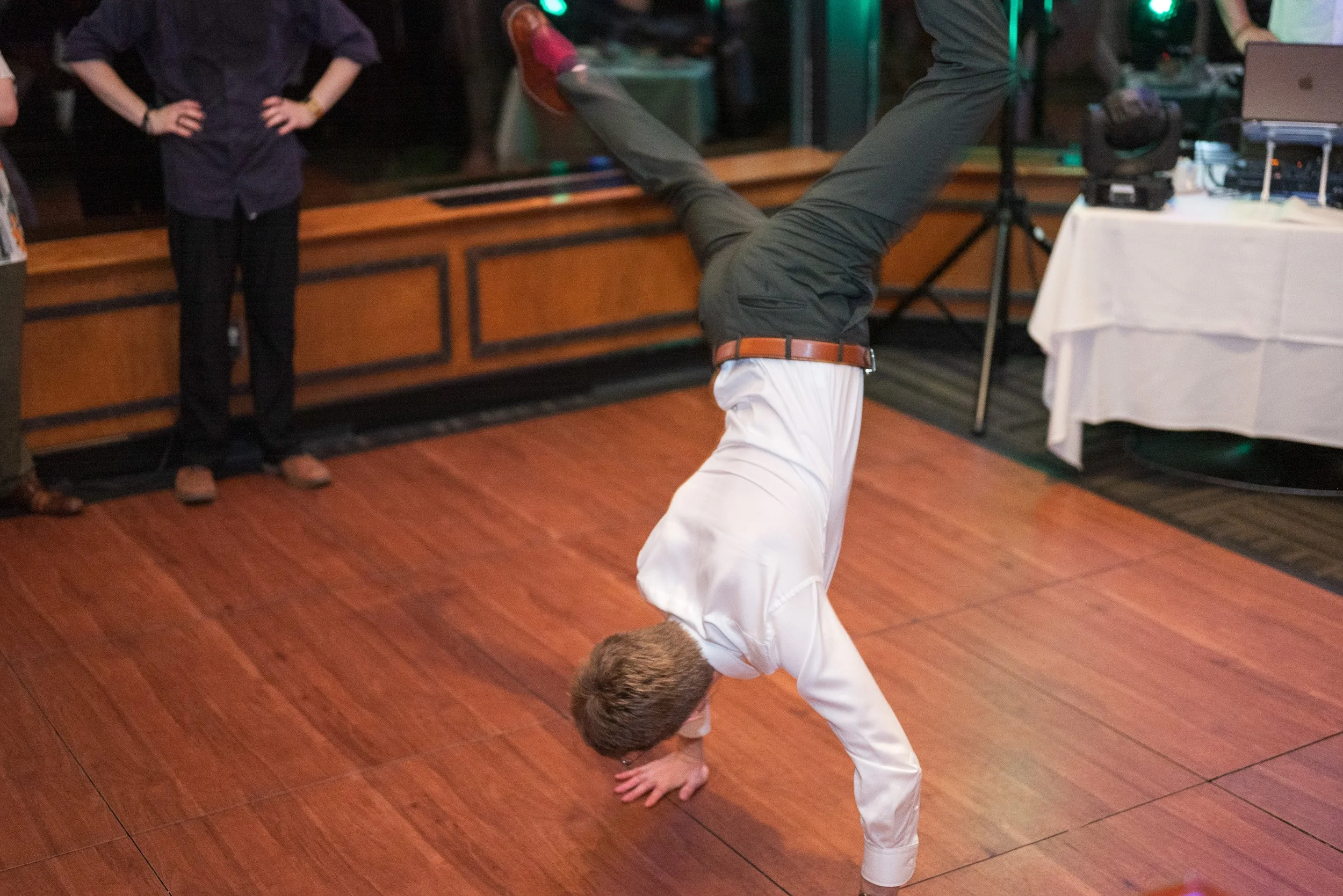 A man performing a handstand on the wooden dance floor at a wedding event, with a few people watching in the background.