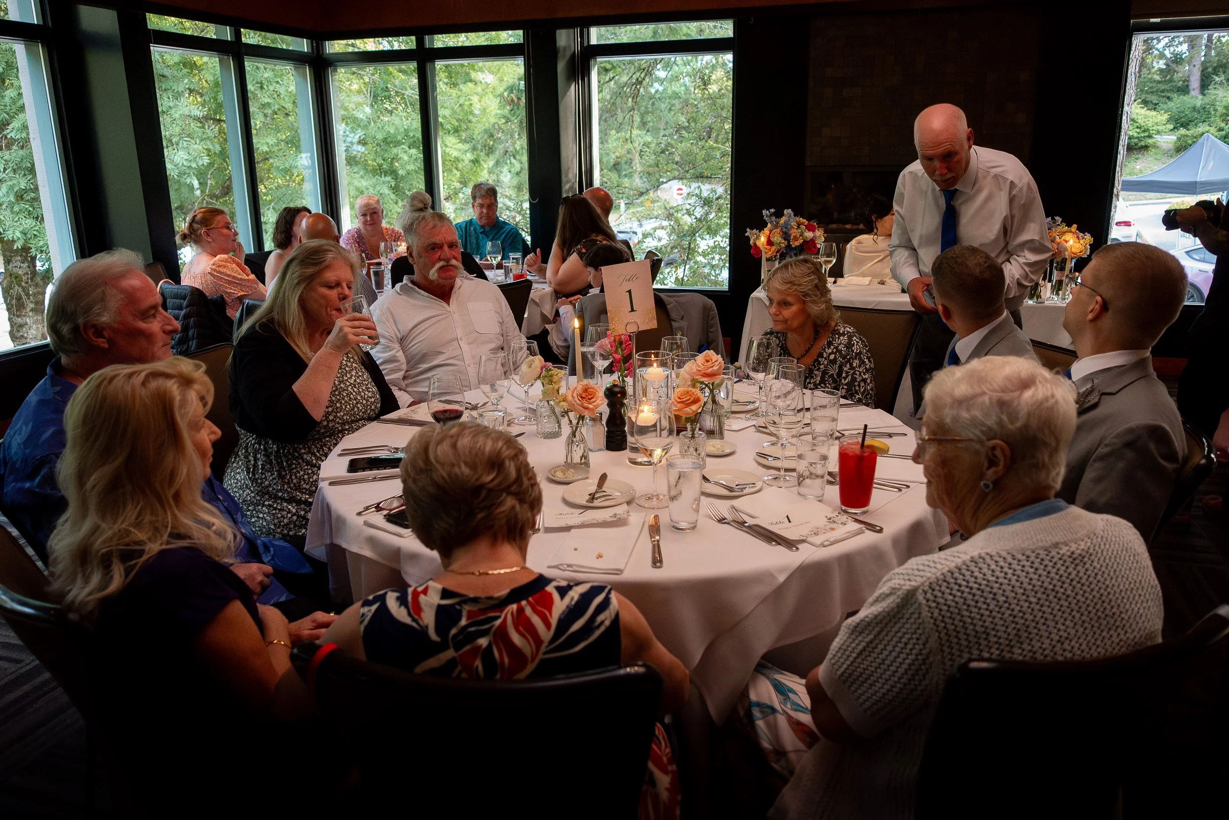 A group of people seated around a round table at a formal event in a restaurant with large windows showing greenery outside. A man in a white shirt is speaking to a woman, and other guests are dining and talking. The table has flowers, glasses, and p
