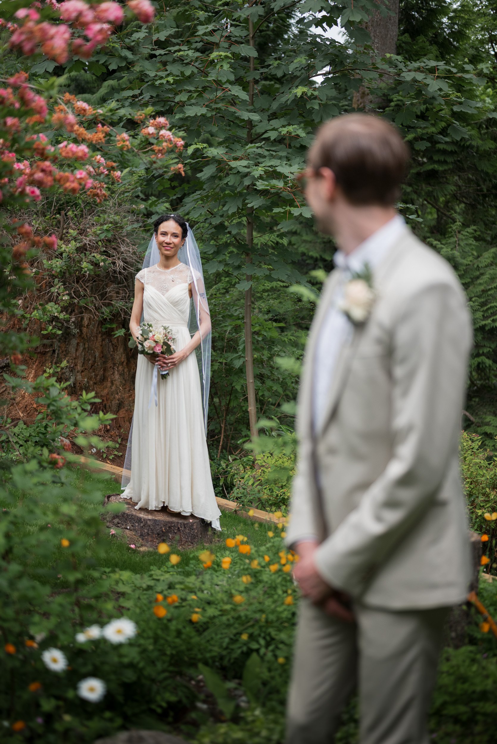 A bride stands amidst greenery and colorful flowers, holding a bouquet, and smiling as she looks at a man in a suit, who is out of focus in the foreground.