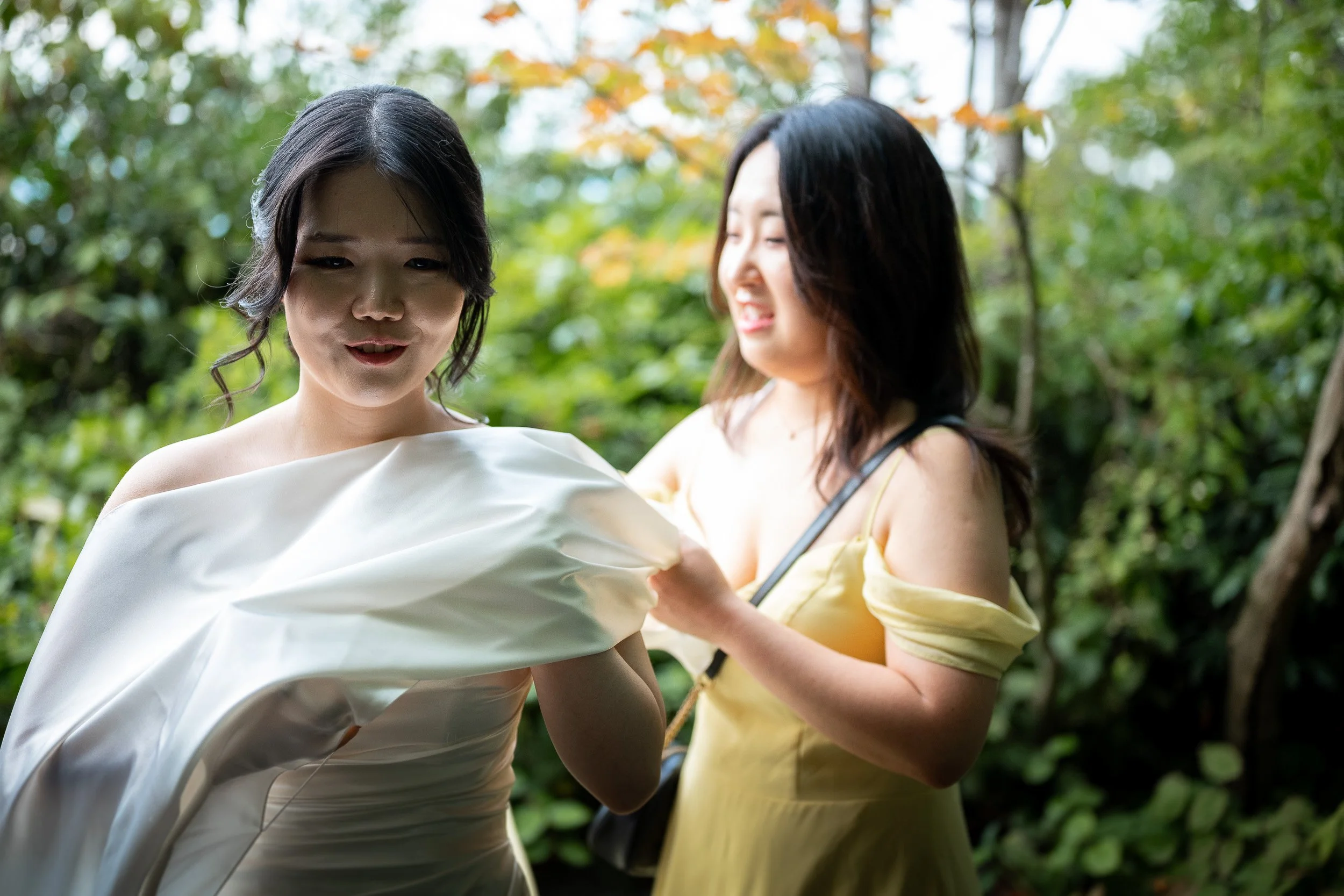 Two women outdoors in a lush, green area, one in a white dress and the other in a yellow dress with thin straps, looking at a piece of fabric or clothing.