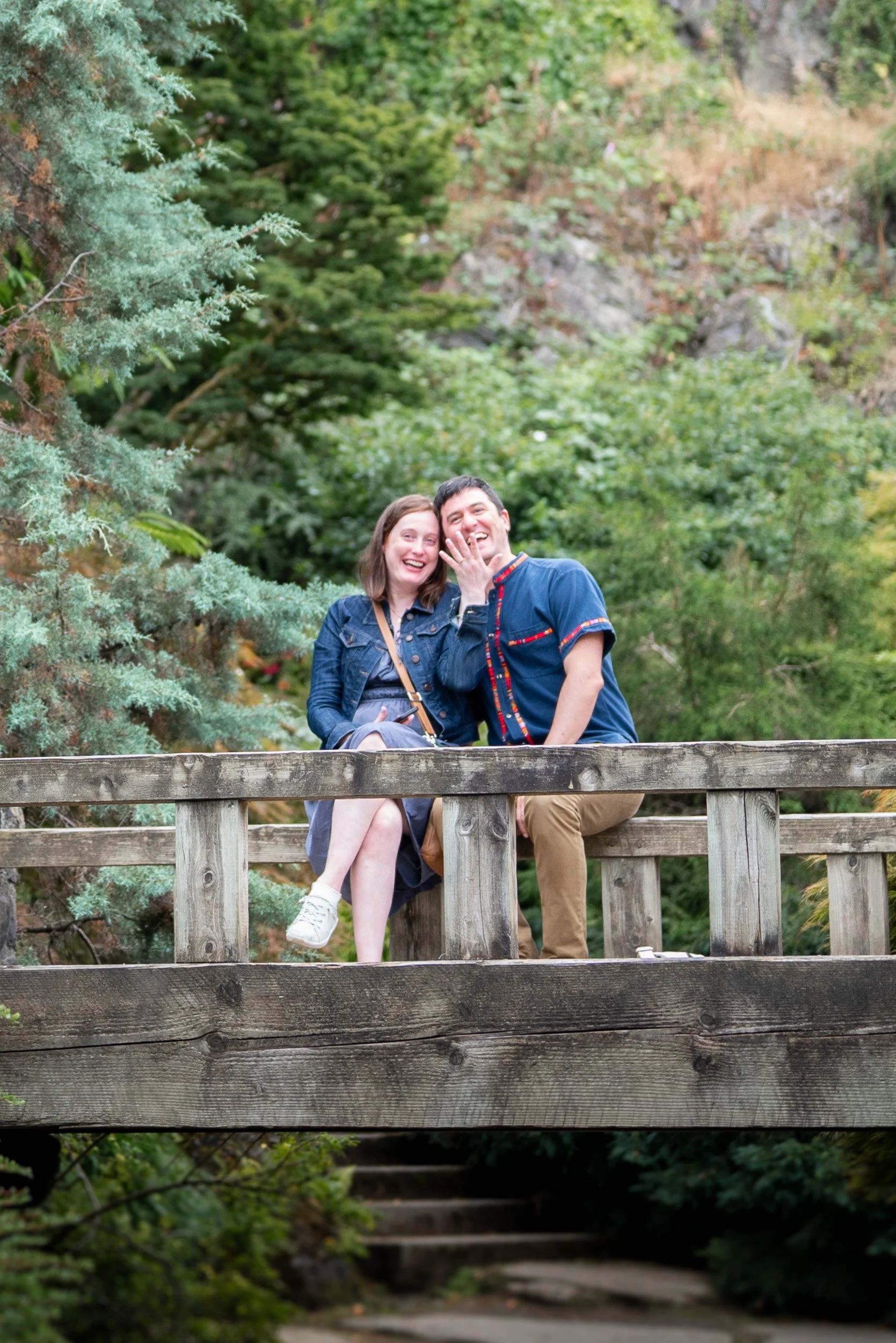 A smiling couple sitting on a wooden bridge surrounded by green trees and foliage.