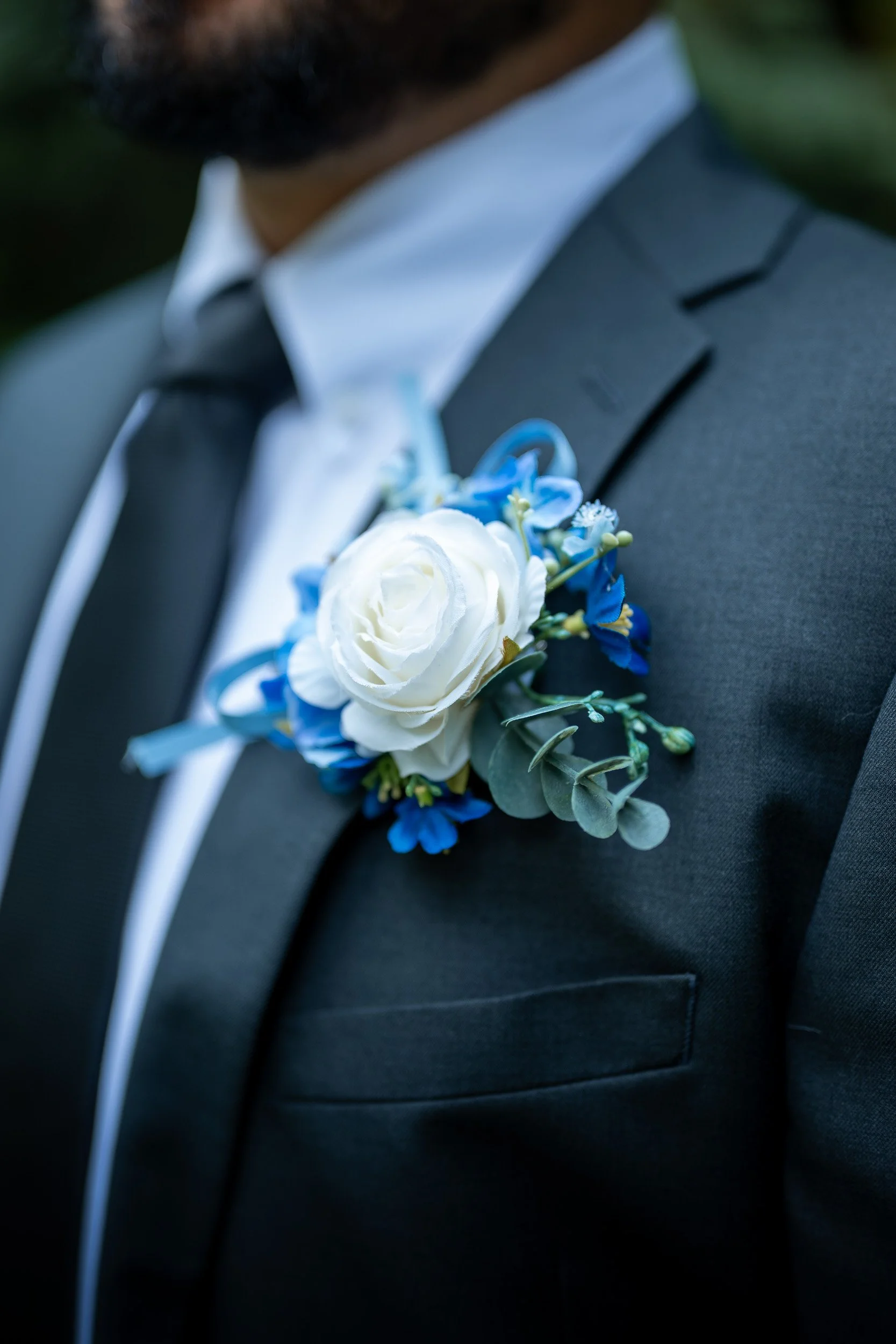 Close-up of a man's suit with a boutonniere made of white rose, blue flowers, and greenery pinned to his lapel, partially showing his beard and shirt collar.