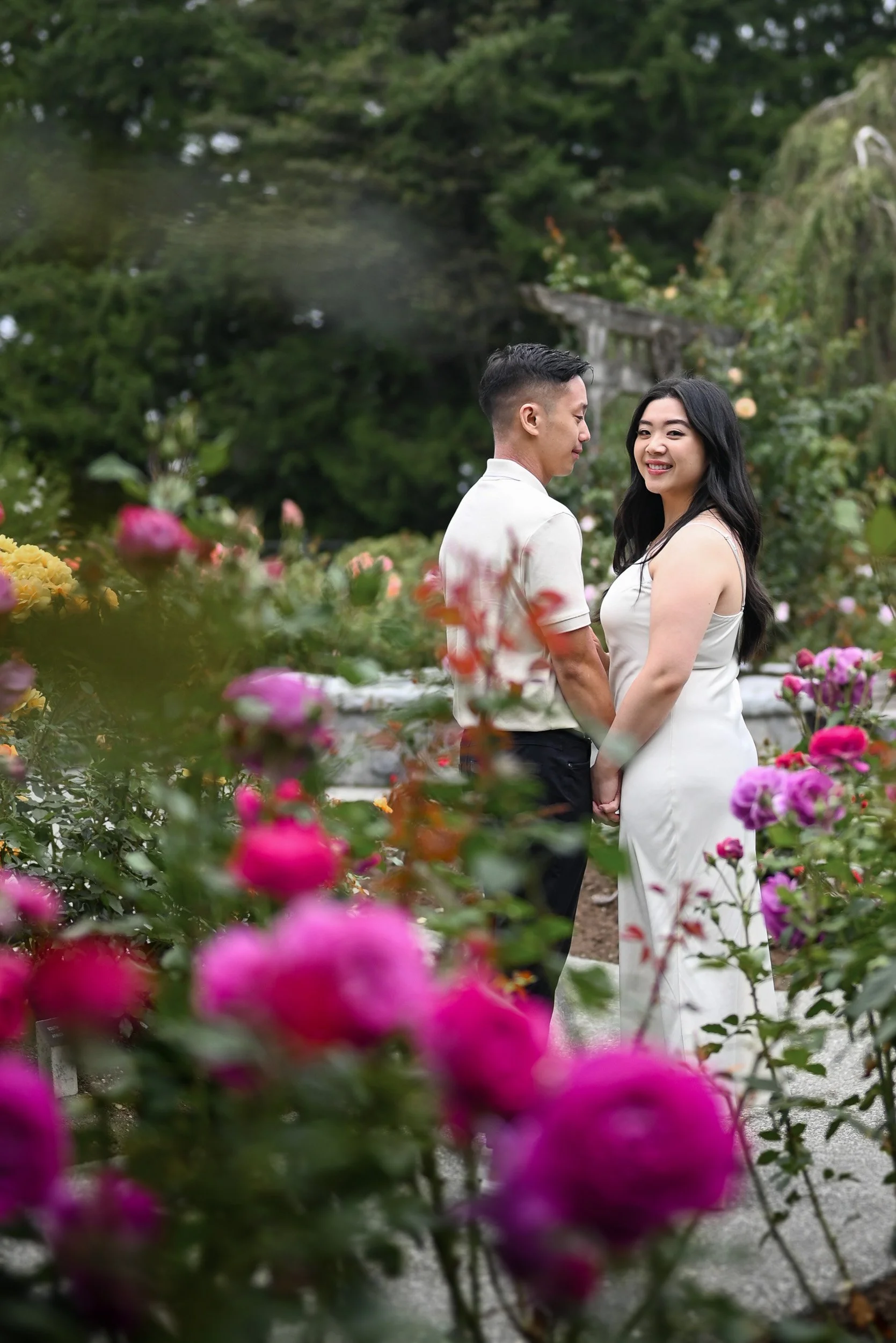 A couple holding hands in a garden surrounded by colorful flowers, with a lush green background and a stone staircase in the distance at Burnaby Mountain.