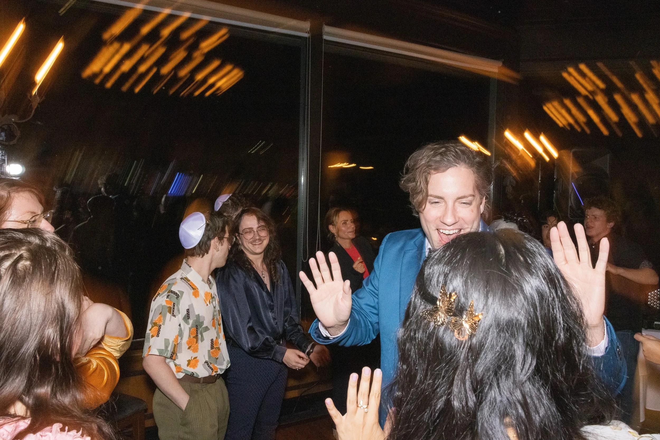 People gathered together at a party or celebration, with one man in a blue suit smiling and raising his hand, surrounded by others chatting and smiling, indoors with a dark background and warm lighting.