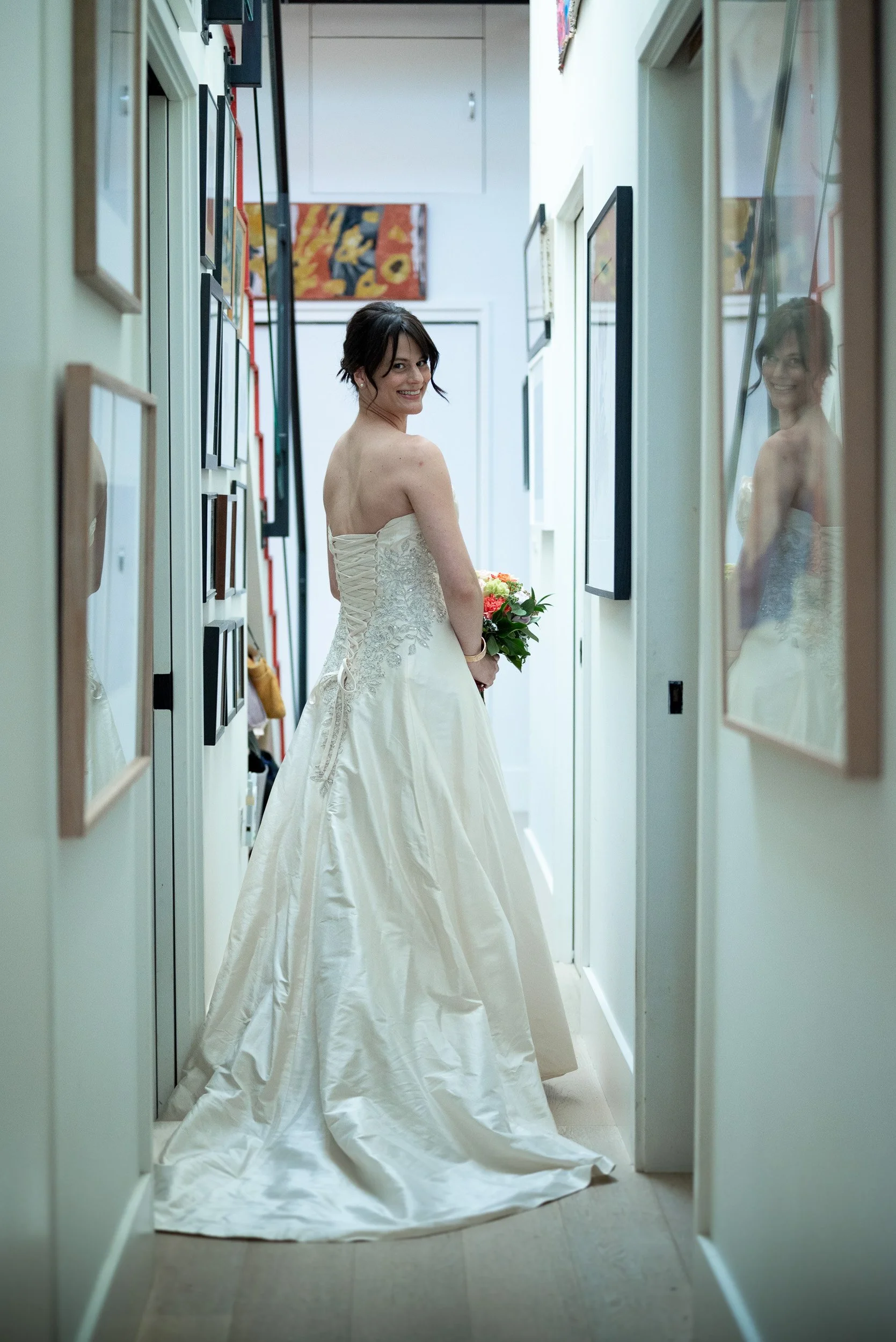 A woman in a wedding dress holding a bouquet, standing in a hallway, smiling at the camera, with her reflection seen in a mirror on the right wall.