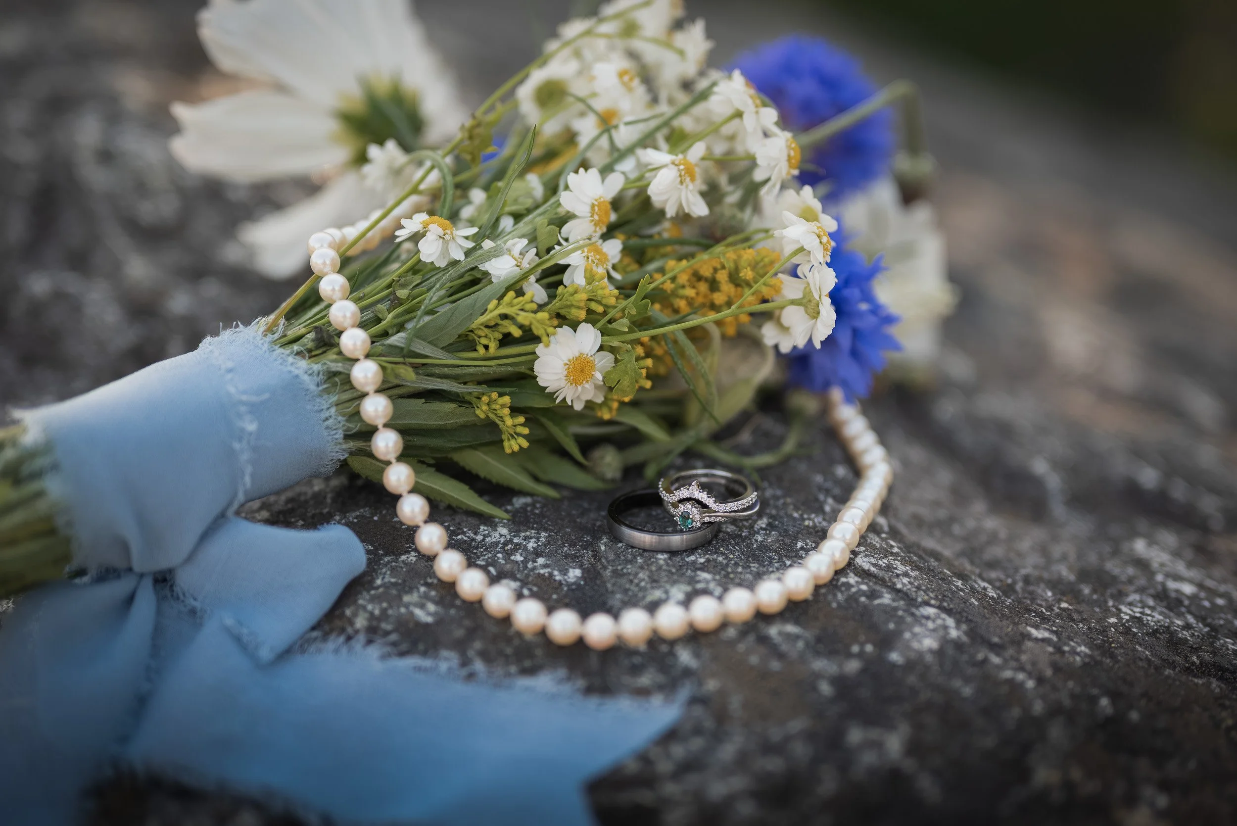 A small bouquet of white daisies, yellow flowers, and blue flowers with a string of pearl necklace, a silver ring with a green gemstone, and a wedding band on a dark granite surface.