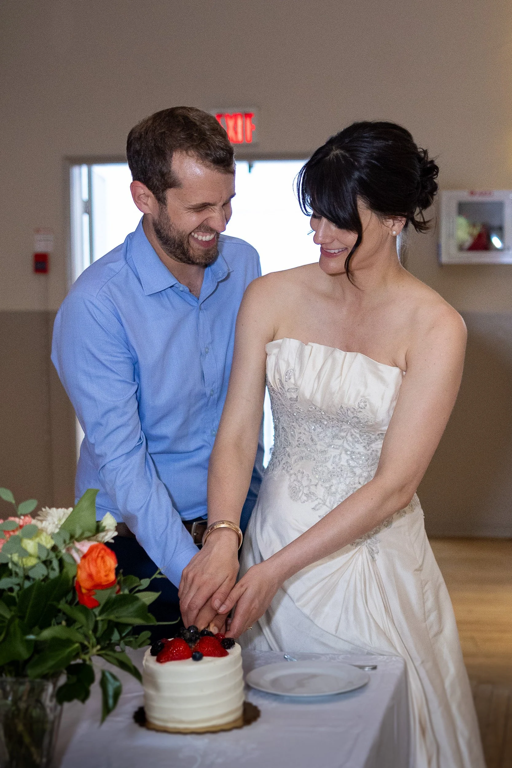 A bride and groom cutting a wedding cake together, smiling and joyful, in a reception hall with flowers and a table.