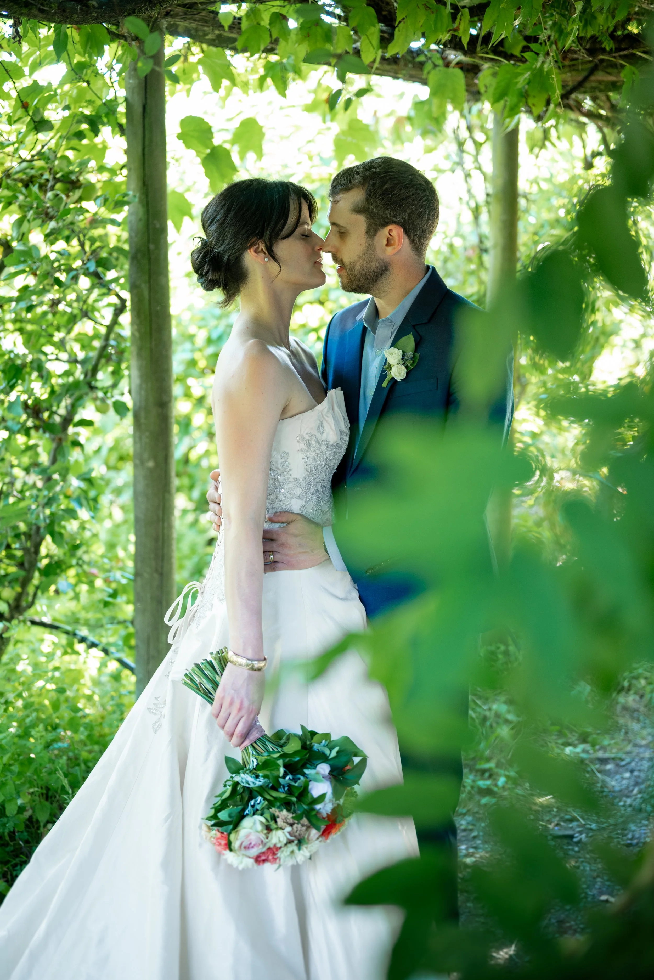A newlywed couple sharing a kiss in a lush green outdoor setting, the woman in a white wedding gown holding a bouquet, and the man in a blue suit with a boutonniere at their wedding in Vancouver, BC