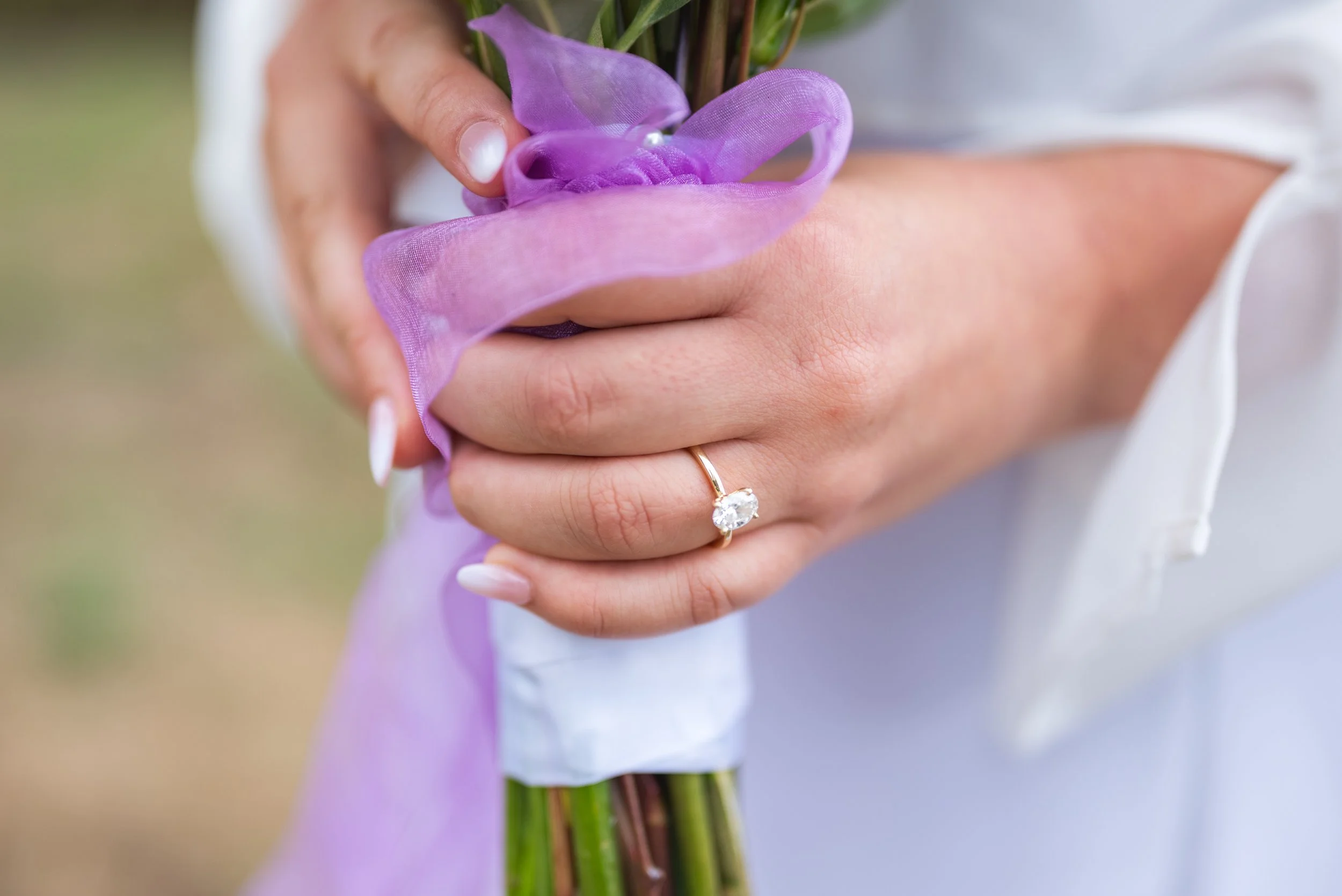 Close-up of a person's left hand holding a small bouquet of purple flowers wrapped with purple sheer fabric, with an engagement ring featuring a large clear gemstone on their ring finger.