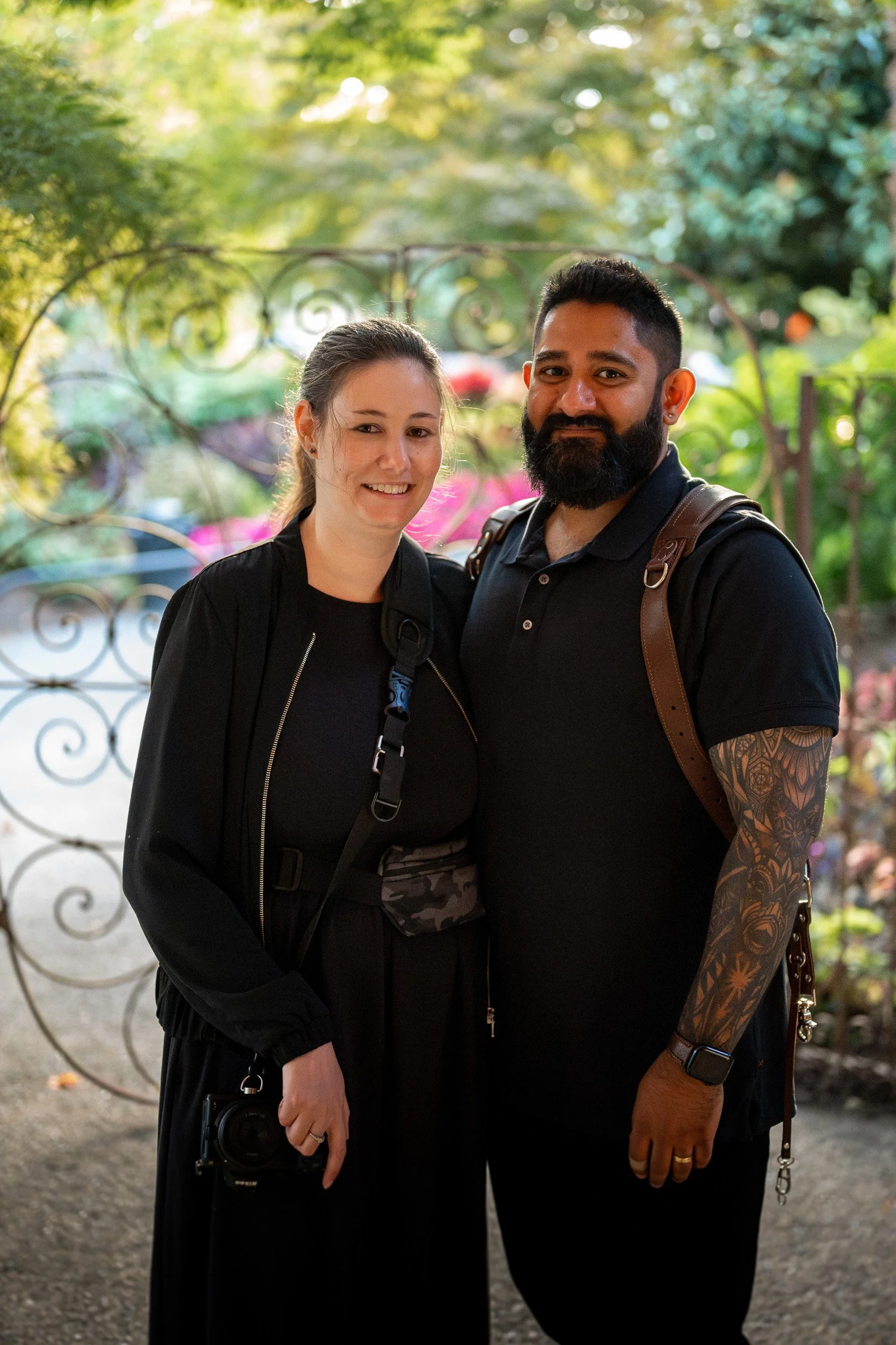 A woman and a man standing outdoors near a wrought iron gate, smiling at the camera surrounded by greenery.