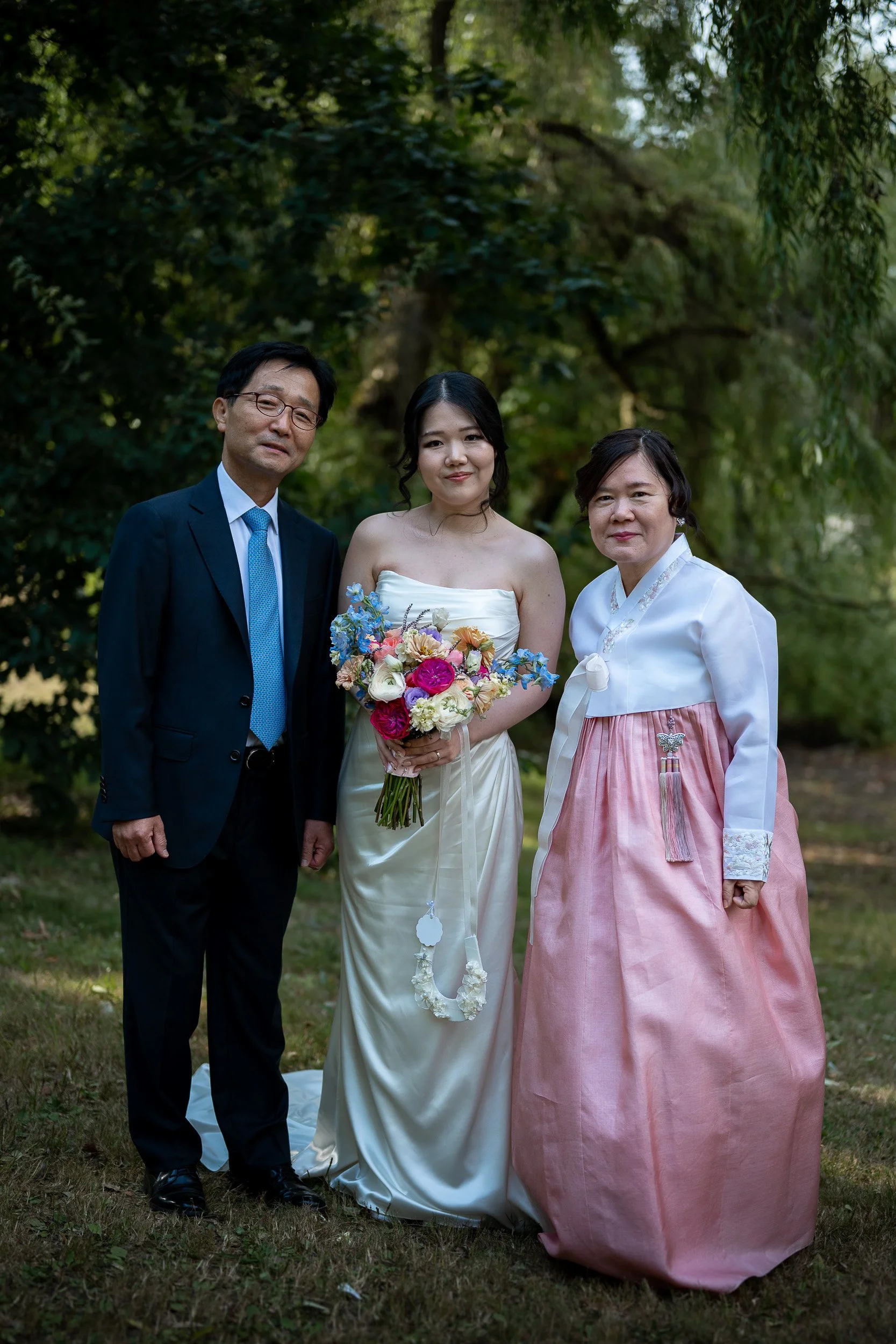 Three people standing outdoors in a wooded area, dressed formally. The woman in the center wears a white strapless dress, holding a bouquet of flowers. The man on the left wears a dark suit with a blue tie and glasses. The woman on the right wears a 