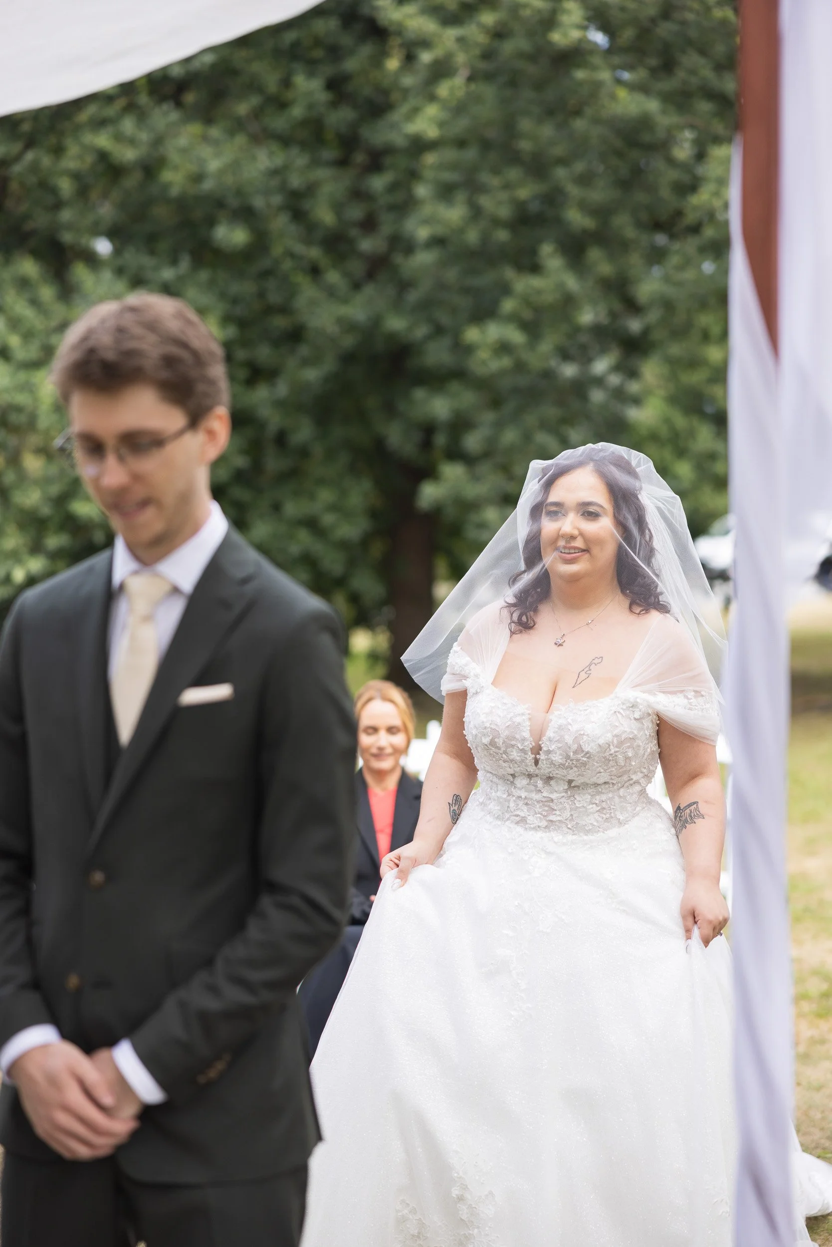 Bride with dark hair in a white wedding gown, smiling, wearing a veil, standing outside during a wedding ceremony, with a groom in a black suit and glasses in the foreground.