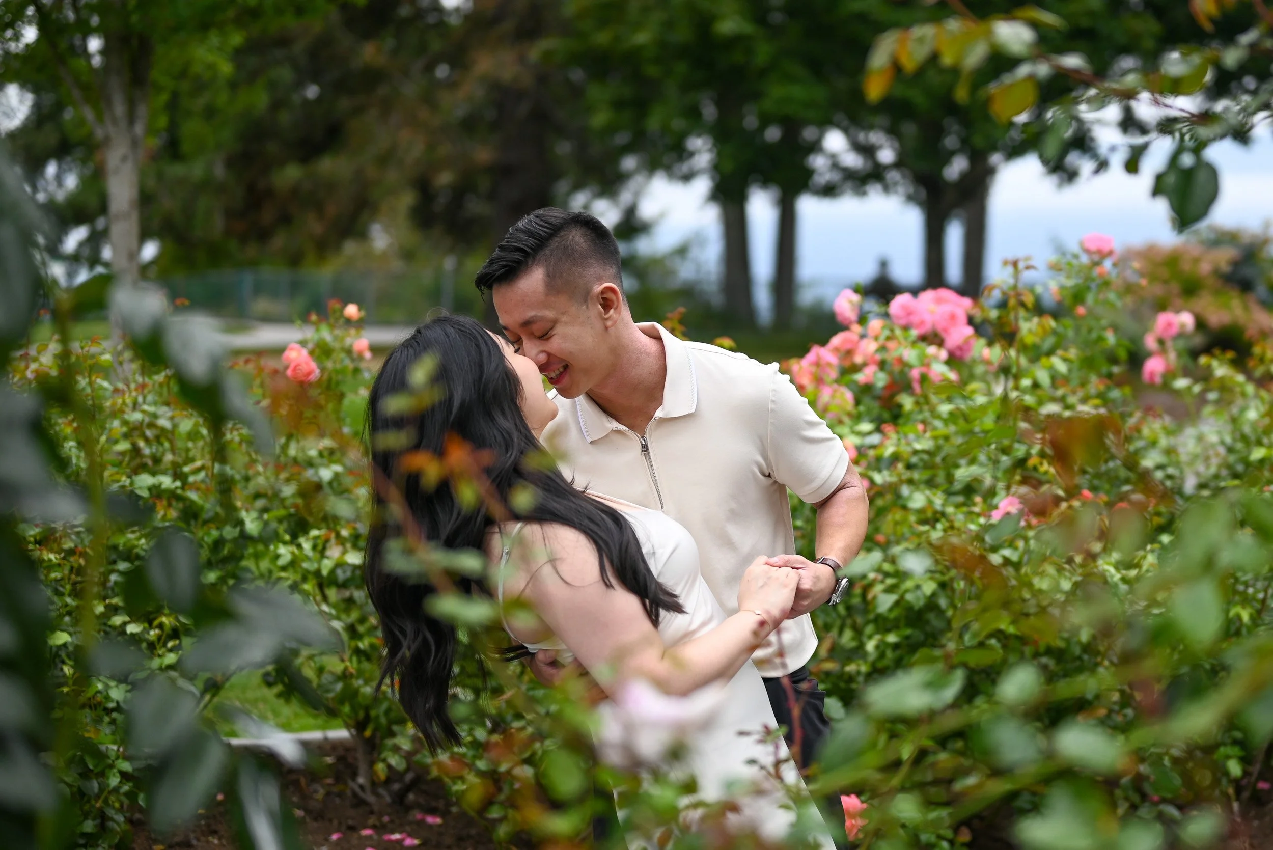 A young couple is standing close together in a garden surrounded by pink roses and green foliage at Burnaby Mountain. They are smiling and appear to be about to kiss at their elopement, with the man gently holding the woman's hands.