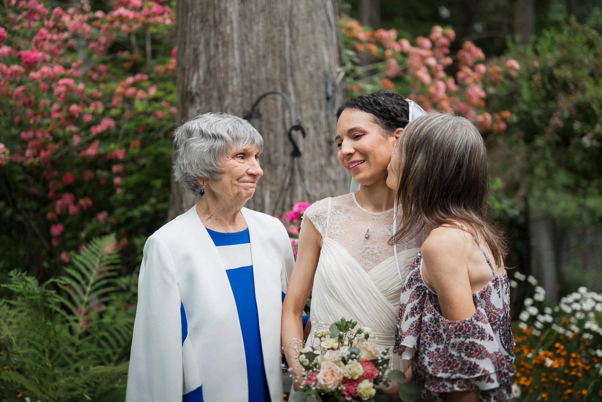 A grandmother, a bride, and two women sharing a moment outdoors during a wedding ceremony surrounded by pink and orange flowers and greenery.