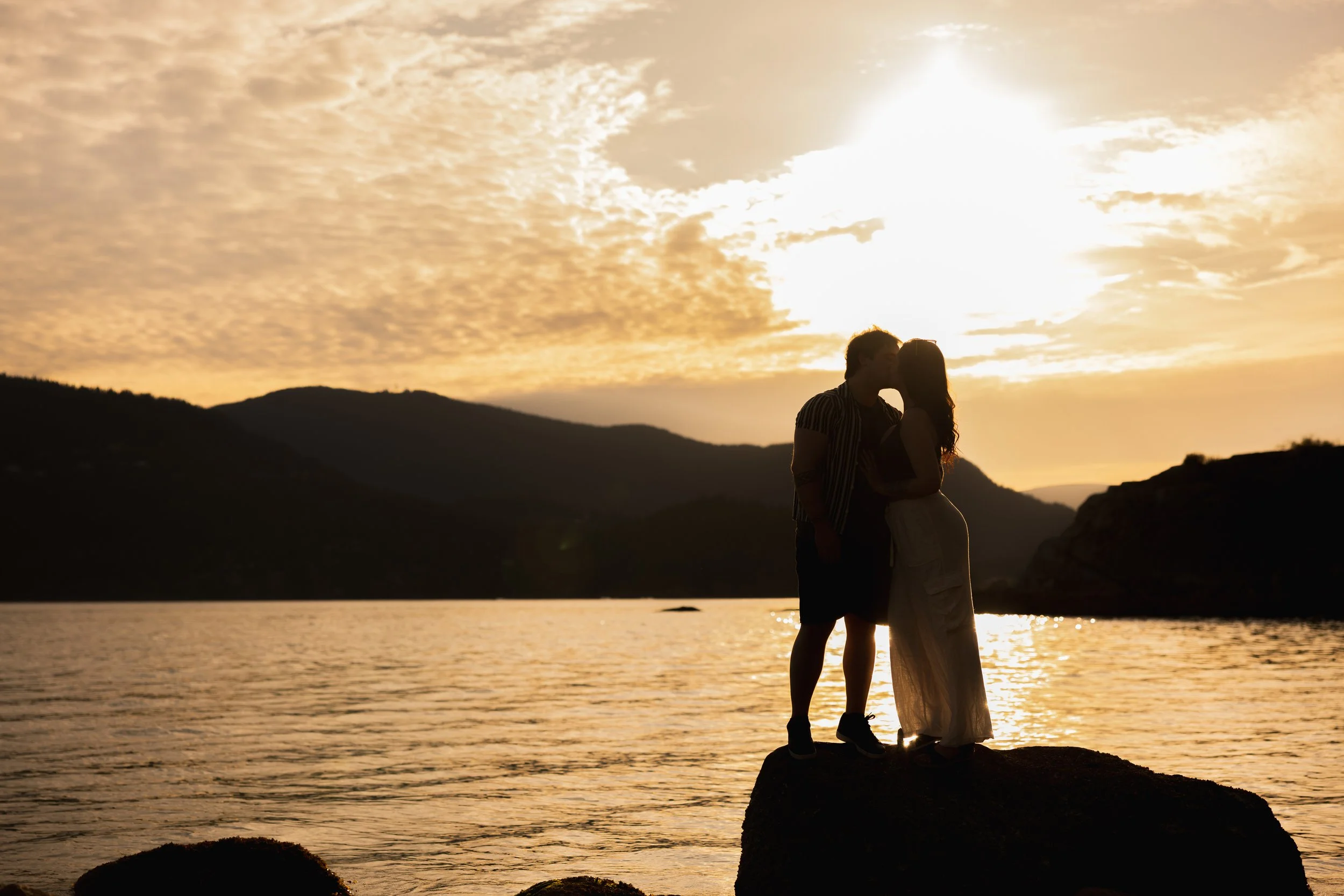 A silhouette of a couple kissing by the water at sunset, with mountains in the background after they got engaged at Whytecliff Park in West Vancouver.